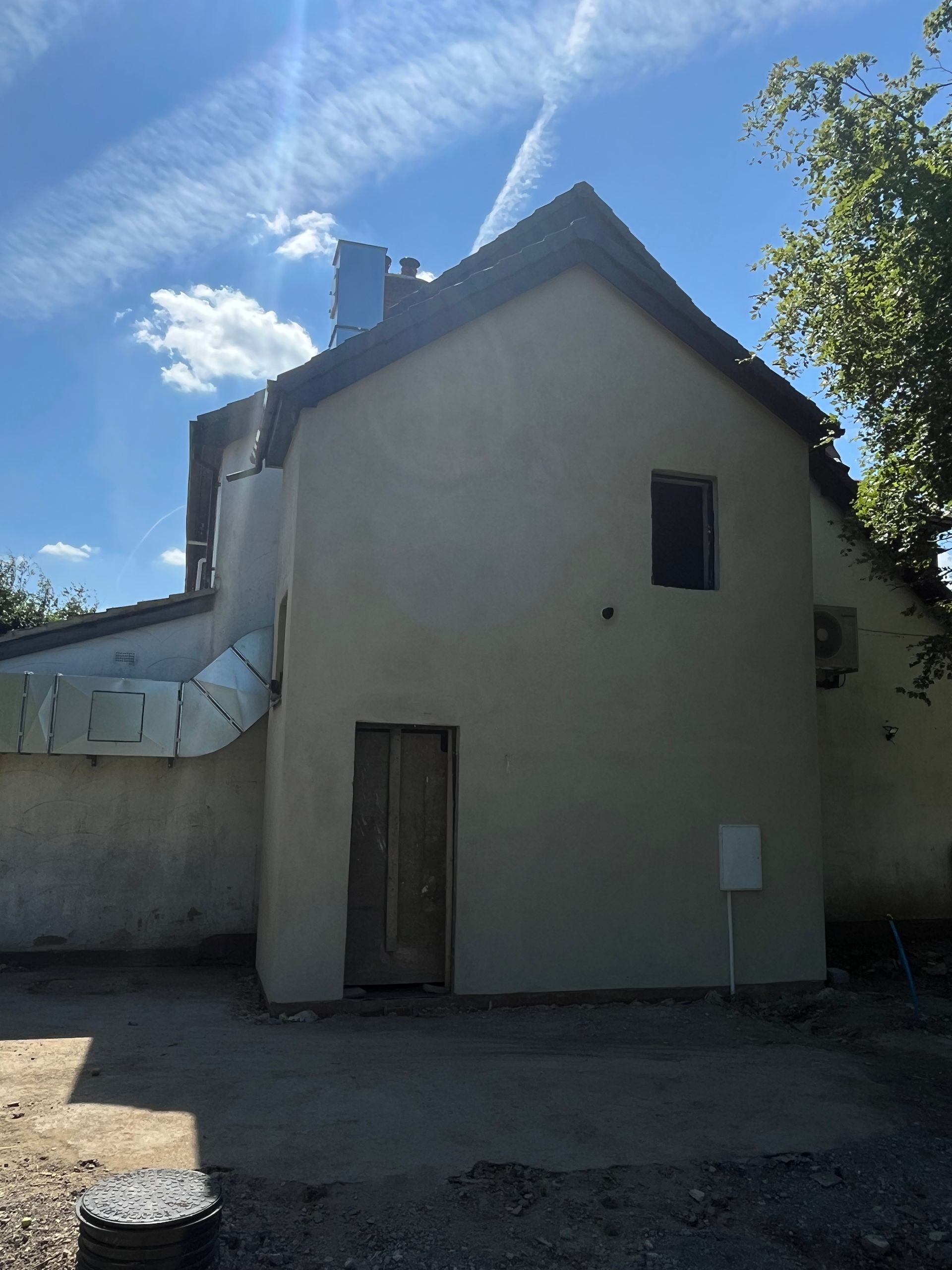 Beige building with a dark door and window, silver chimney, and blue sky with wispy clouds.