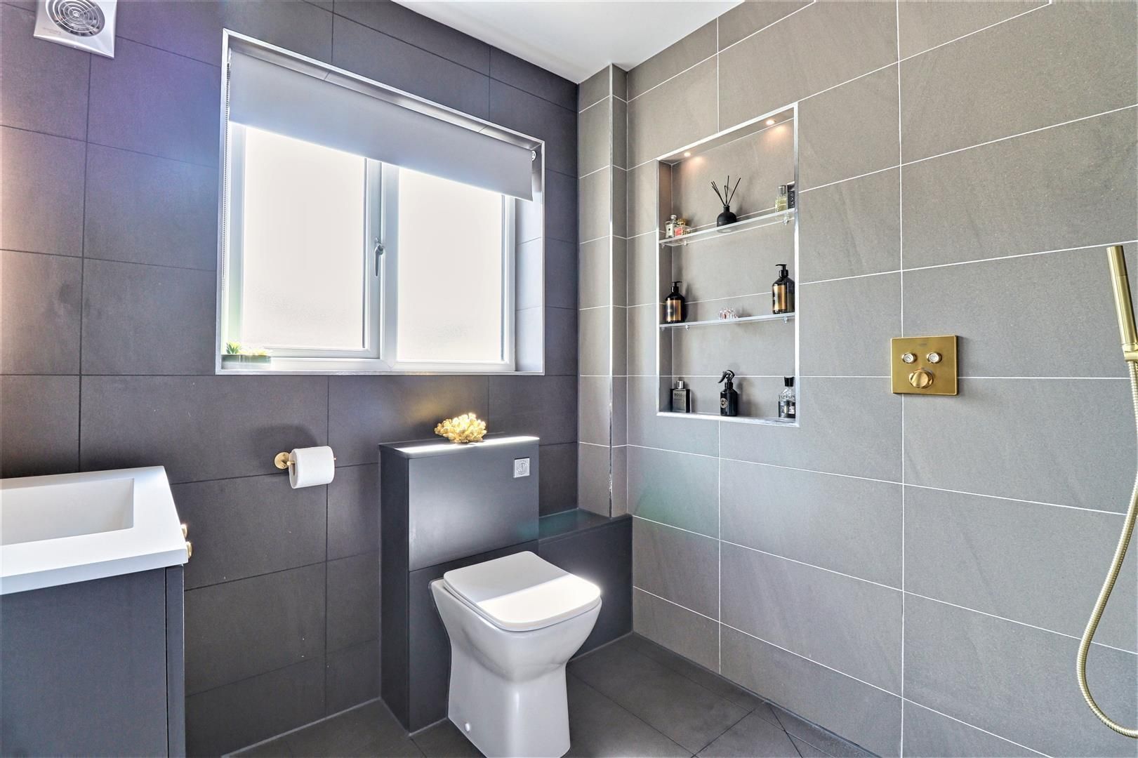 Modern gray bathroom with a window, toilet, and built-in shelving. Gold fixtures and square tiles.