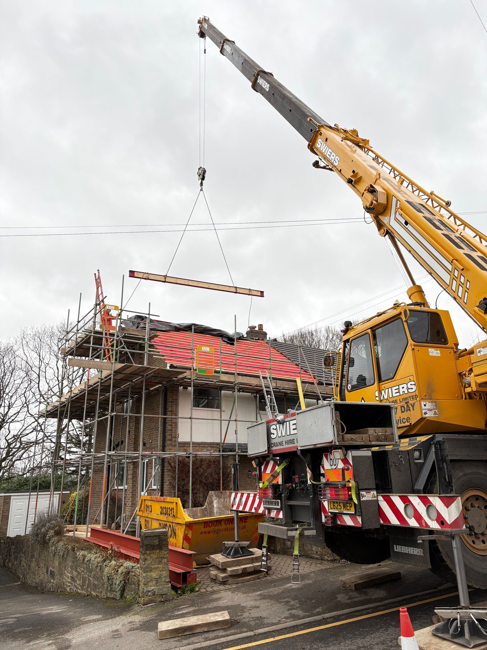 A crane lifting a beam onto a house under construction. Yellow crane, scaffolding, red roof.