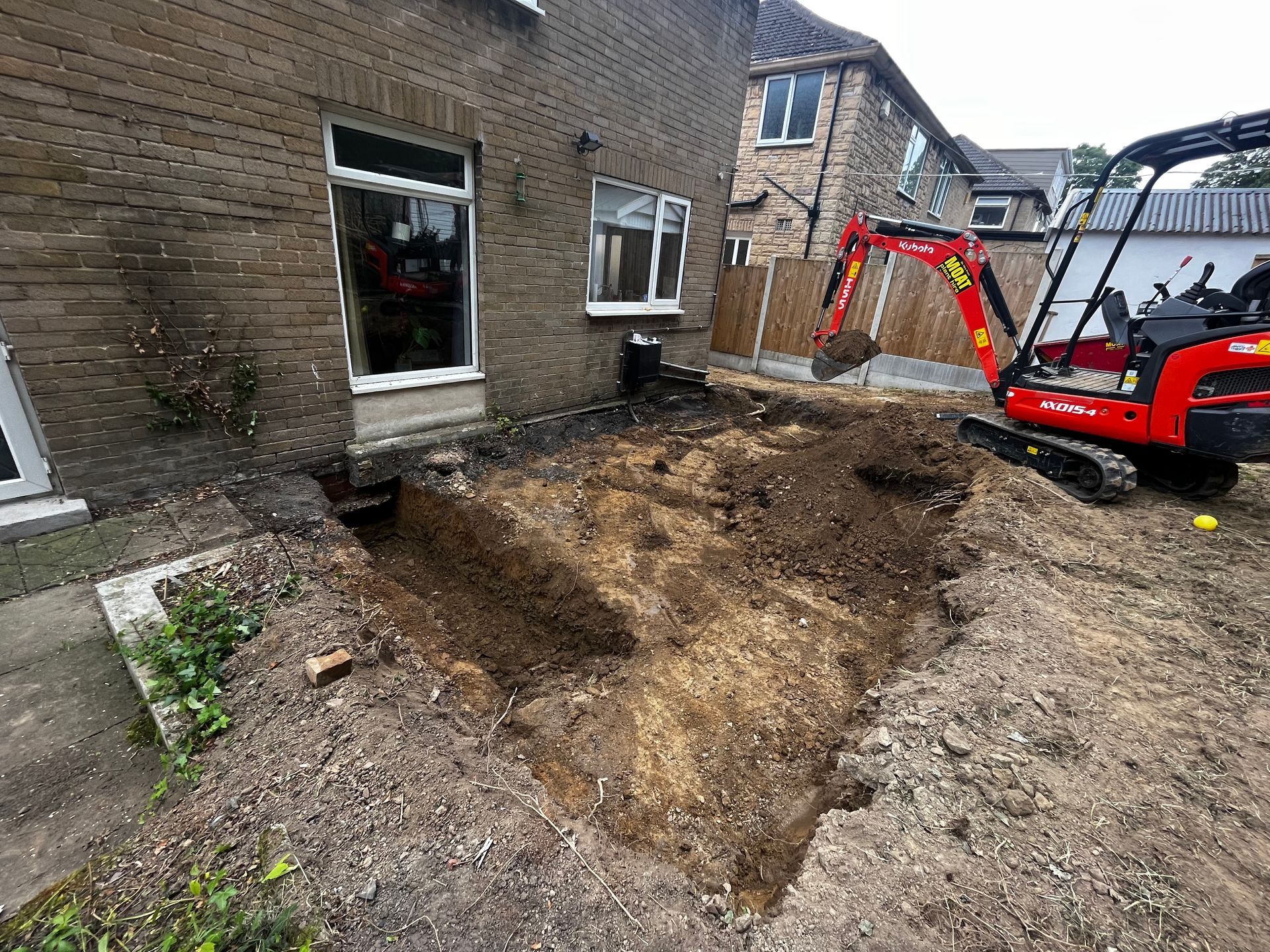 A small red excavator digs in a dirt patch next to a brick building.