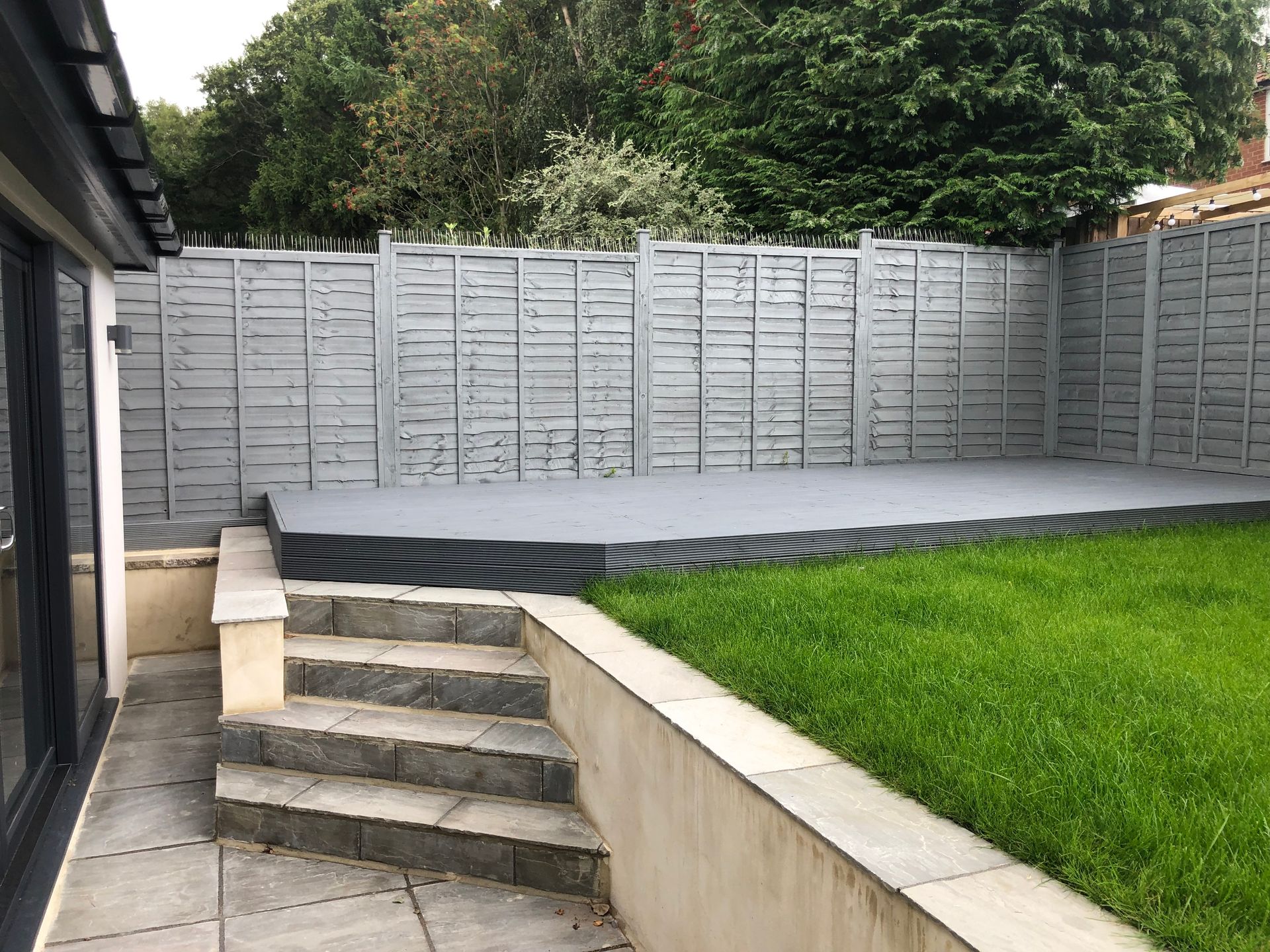 Backyard patio with steps leading to a raised platform; gray painted fence and deck; green grass.