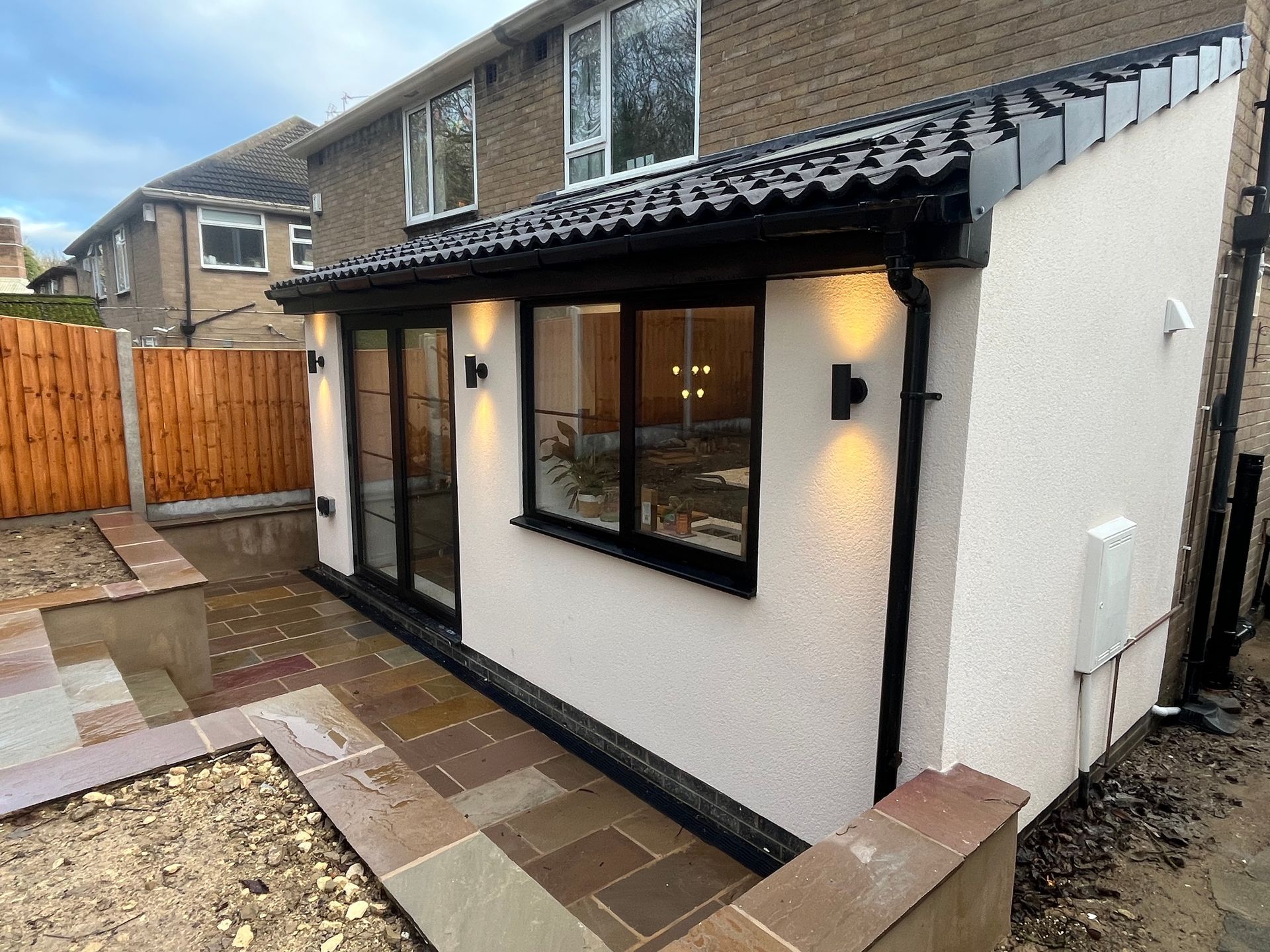 Newly built extension with black-framed windows, black gutters, and a cream exterior, adjacent to a paved patio area.