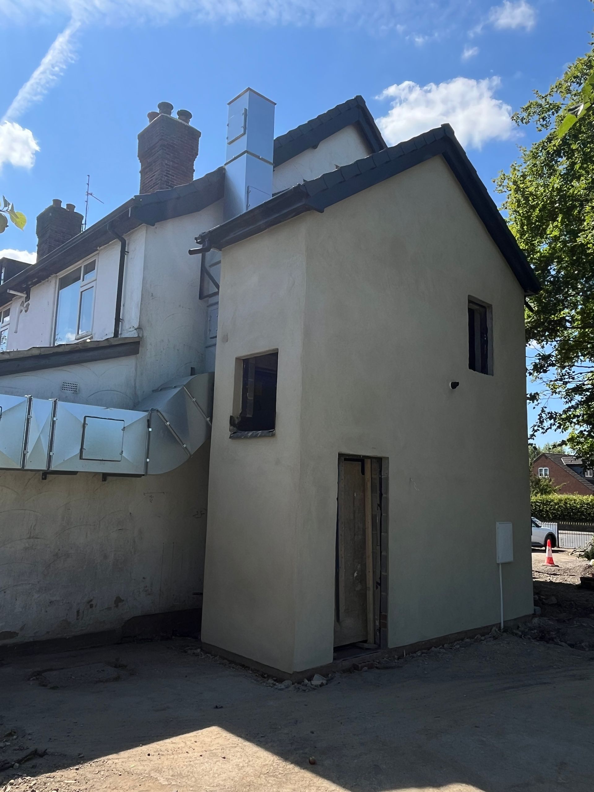 Exterior view of a light-colored building addition; small square windows, wooden door, metal exhaust pipe.