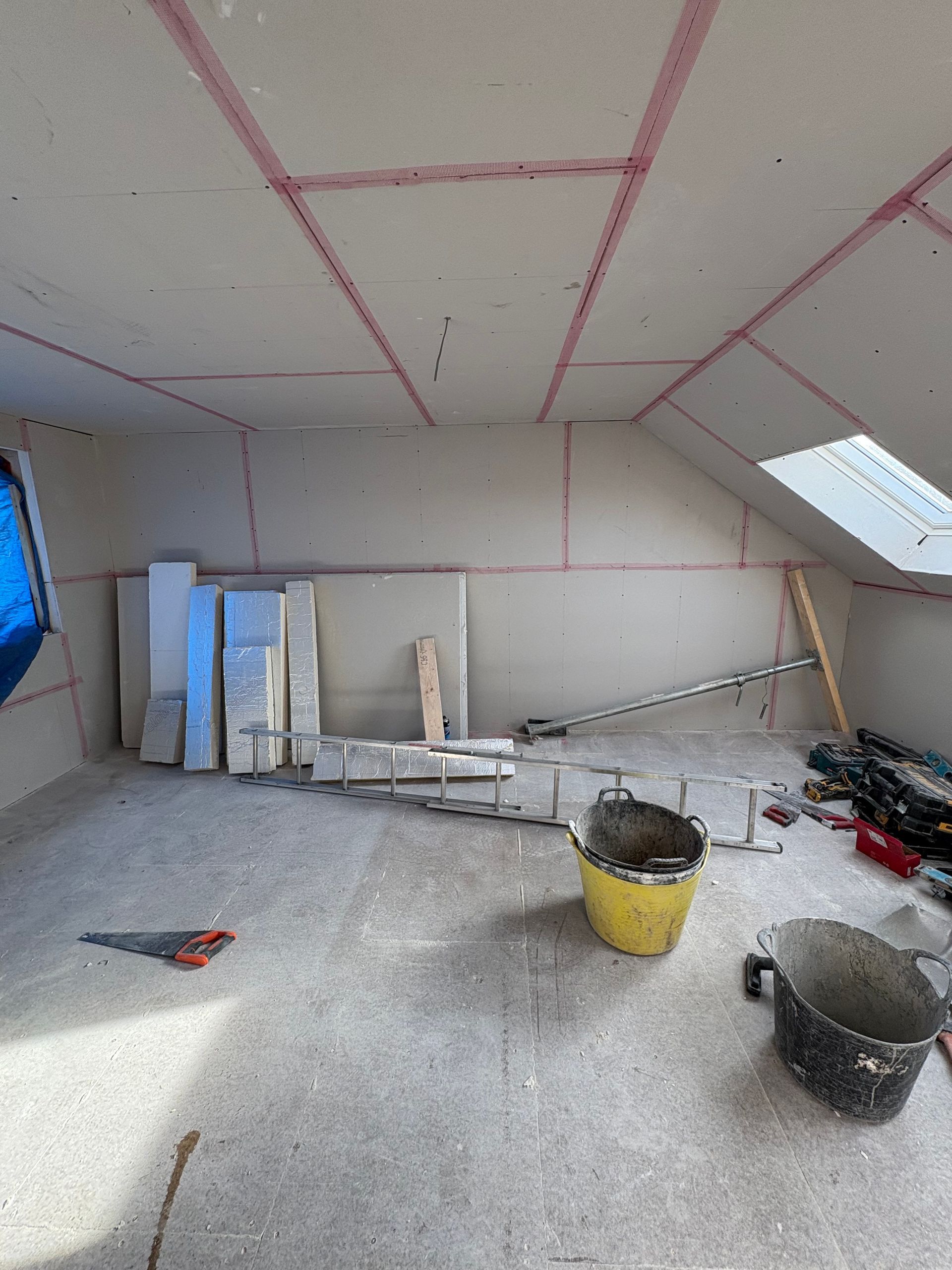 Interior room under construction, drywall installed. Red lines mark seams. Tools, buckets, and materials are scattered.