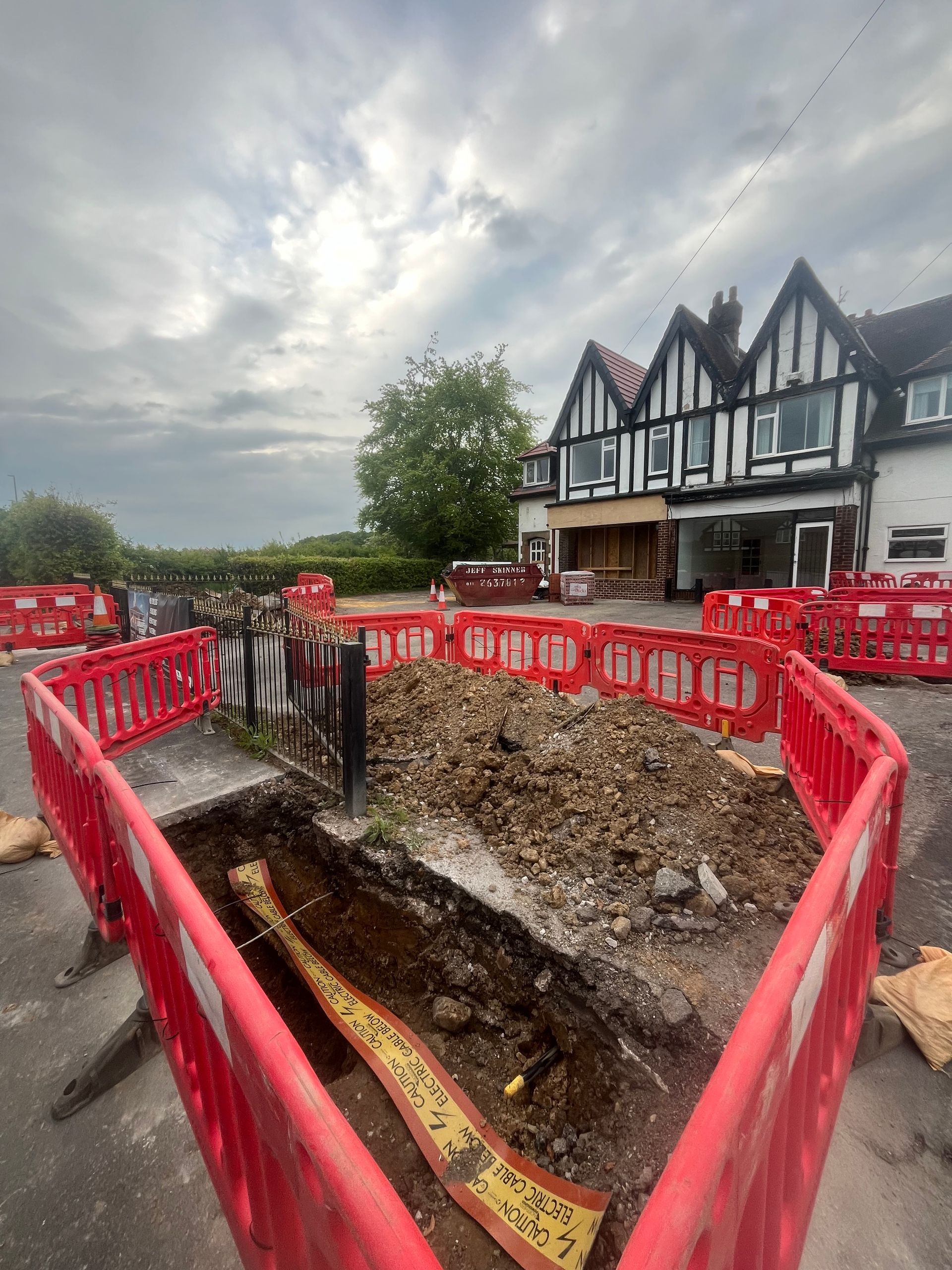 Trench dug in asphalt with red barriers in front of a Tudor-style building.