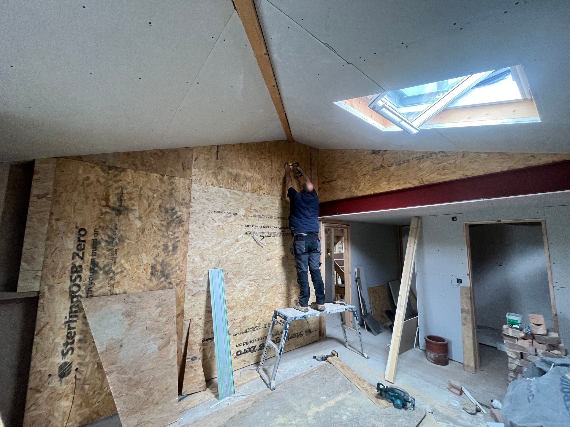 A person on a step stool installing wall panels in a room under construction. Skylight visible above.
