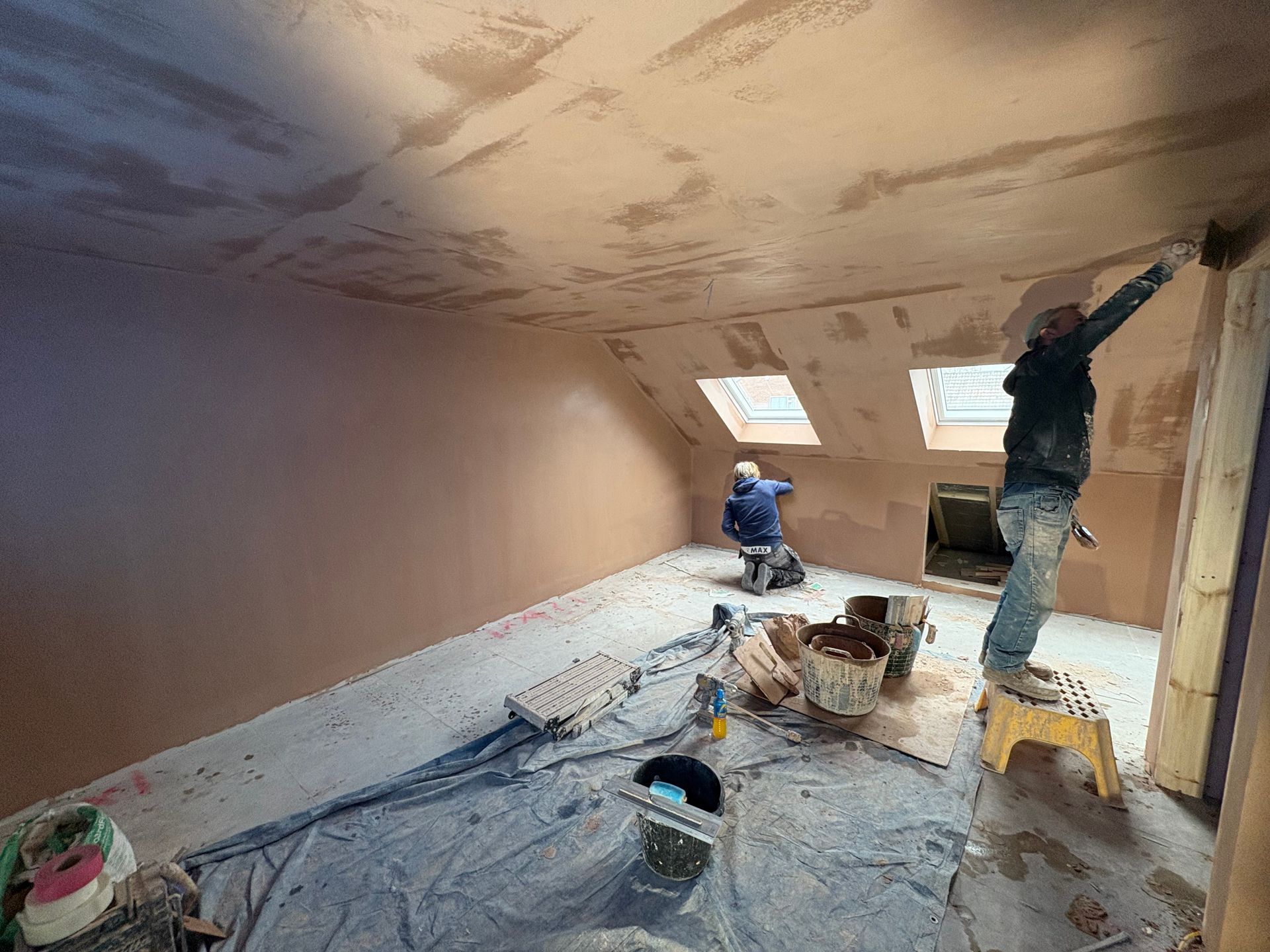 Two plasterers working on an interior room with slanted ceiling, applying plaster; brown walls and ceiling
