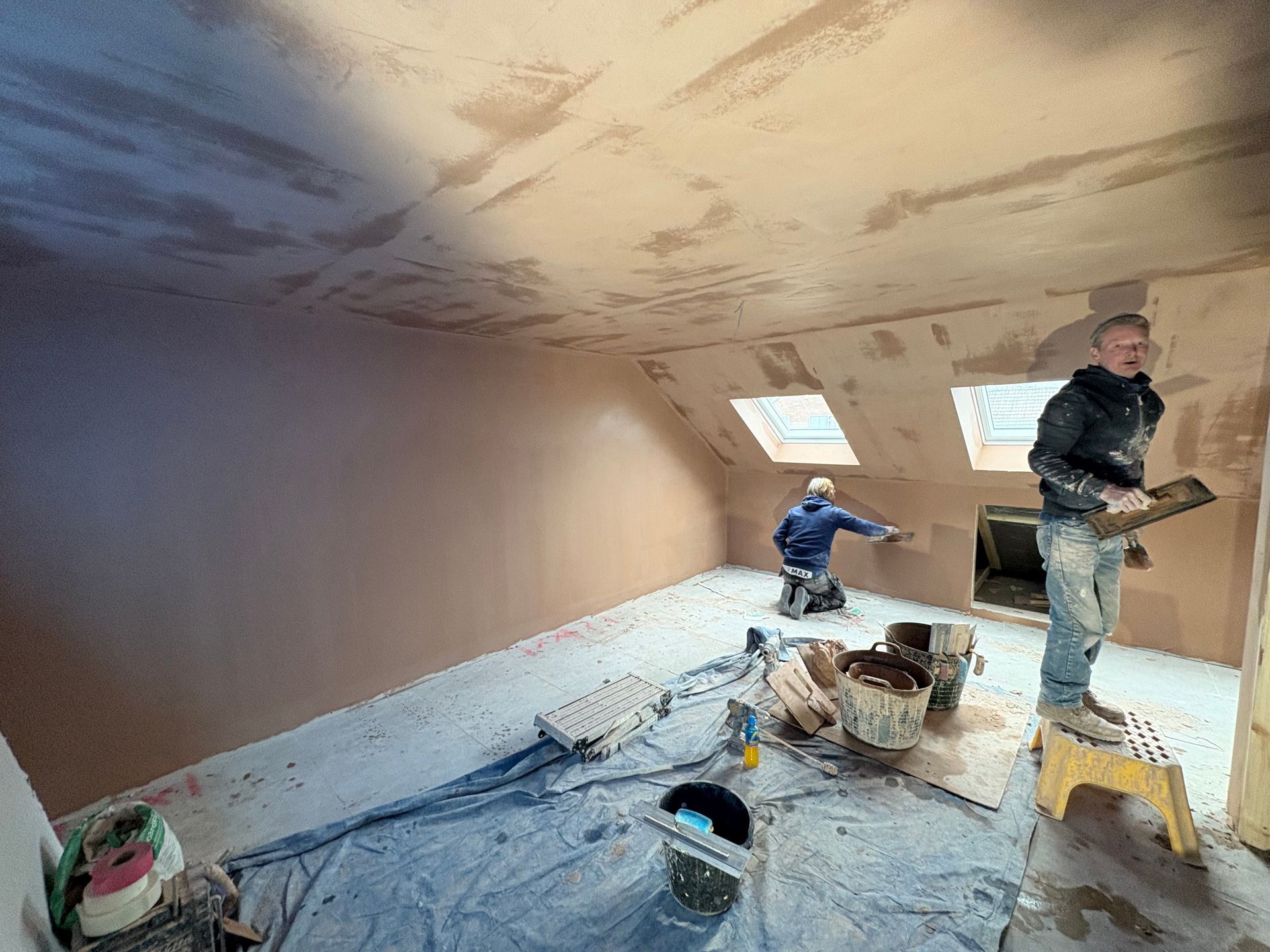 Two people plastering an attic room with pinkish plaster. One stands on a stepstool.