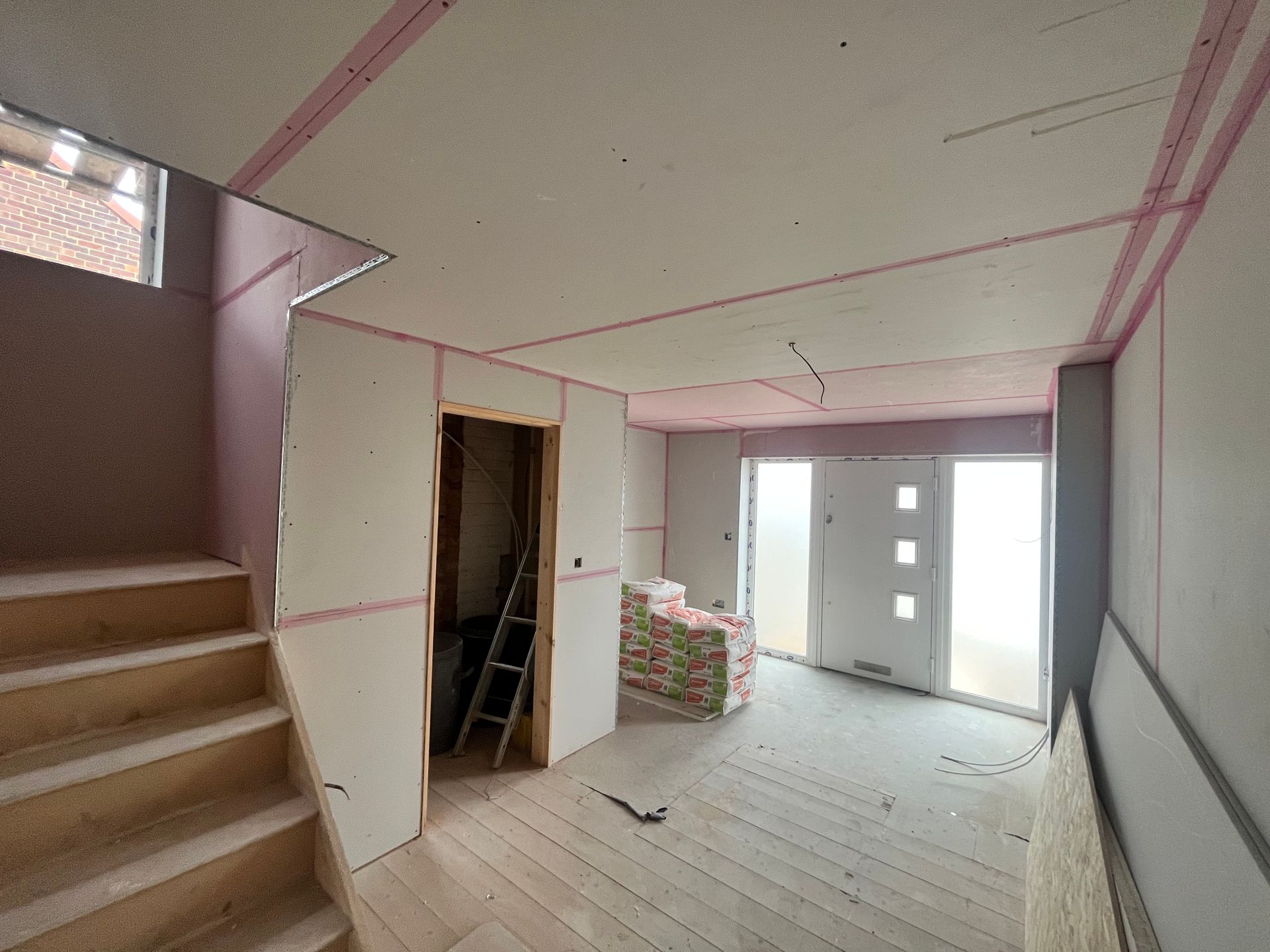 Interior of a home under construction with drywall installed; stairs, door frames, and stacks of building materials visible.