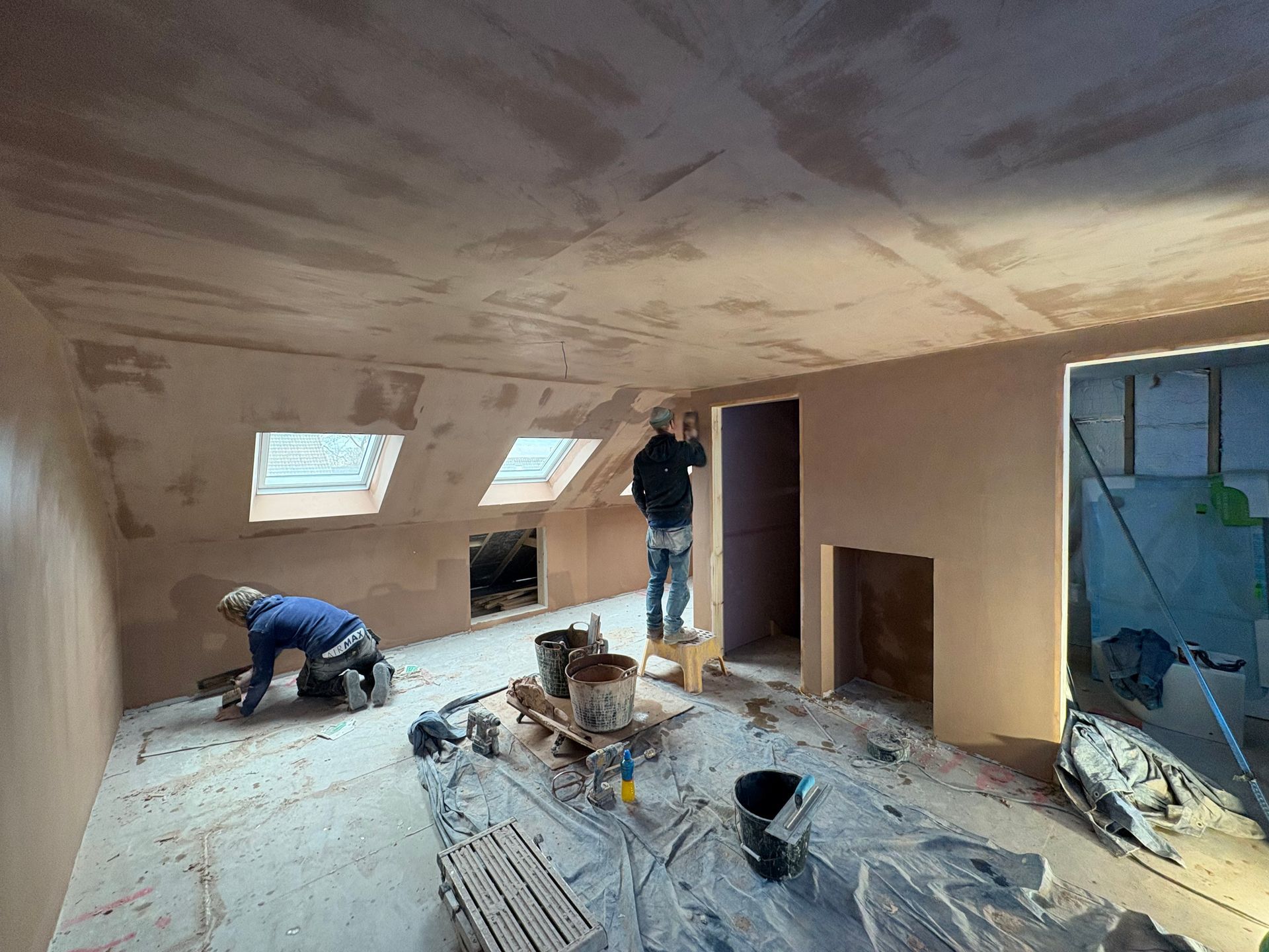 Two people plastering an attic room. One kneeling, the other on a stool. Light pink walls and ceiling.