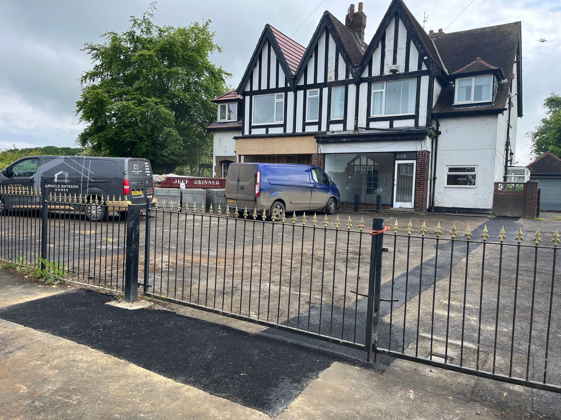 Tudor-style building with vans parked in front, black fence. Cloudy sky.
