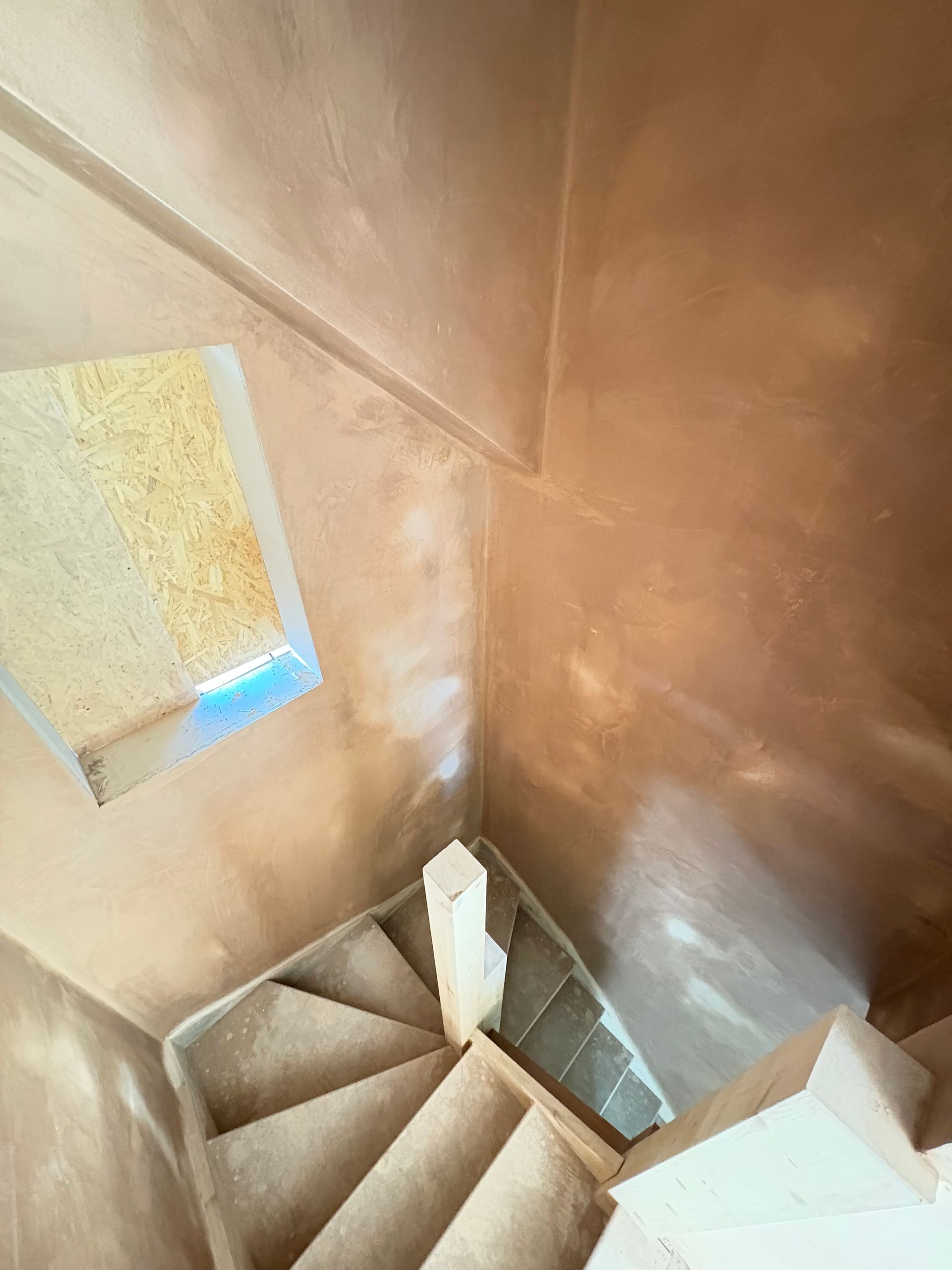 Looking down a newly plastered staircase, light from a window illuminates the walls and steps.