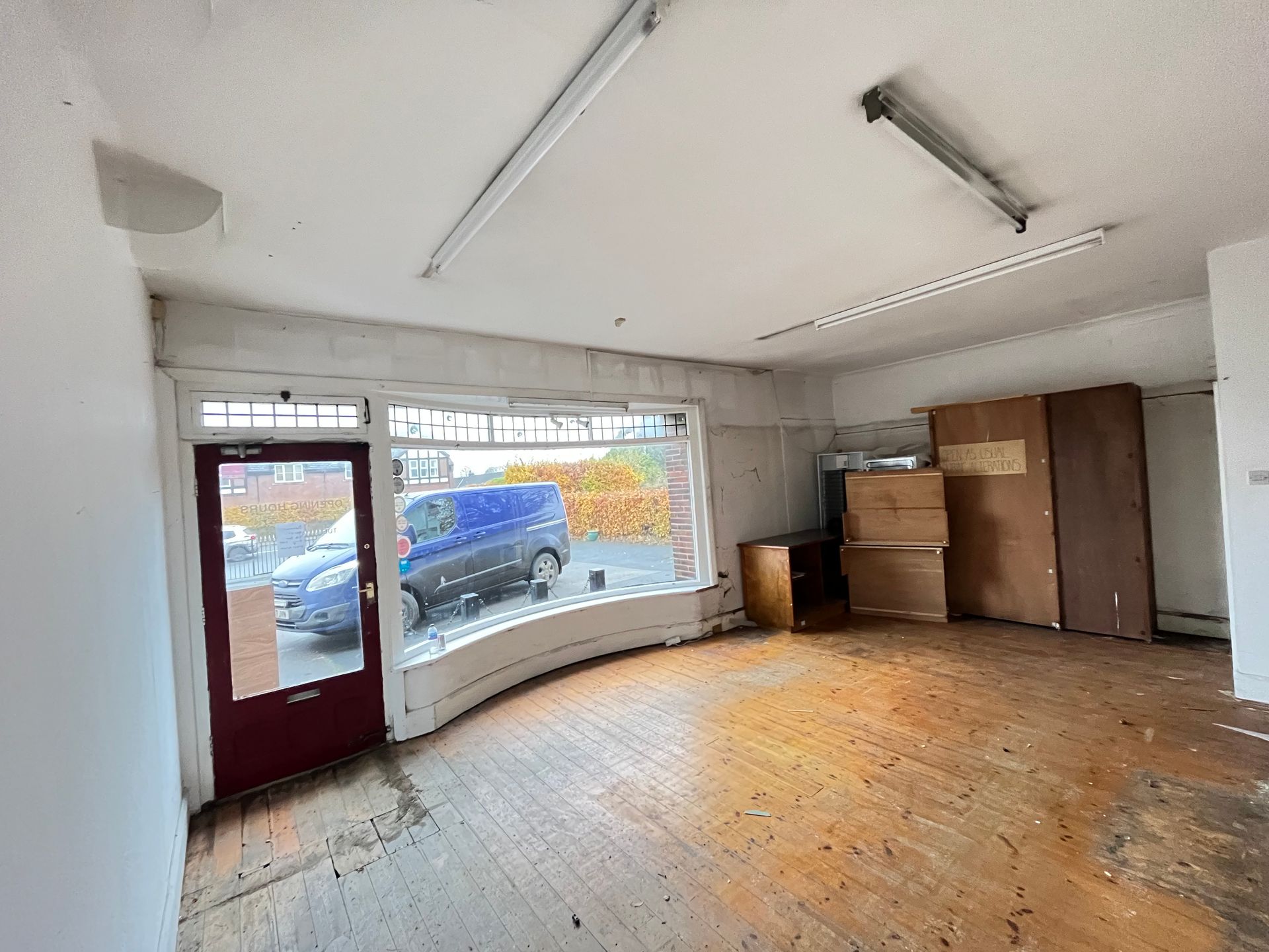 Empty commercial space with a curved storefront window, bare wood floor, and a few cardboard boxes.