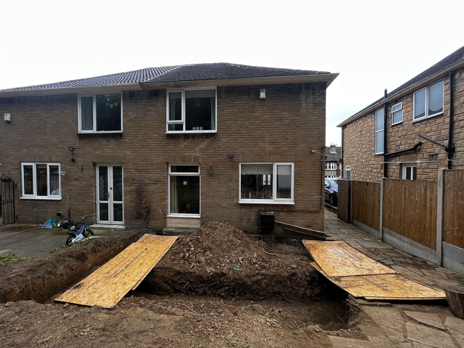 Backyard construction site: house, dug trench, plywood walkways, pile of dirt.