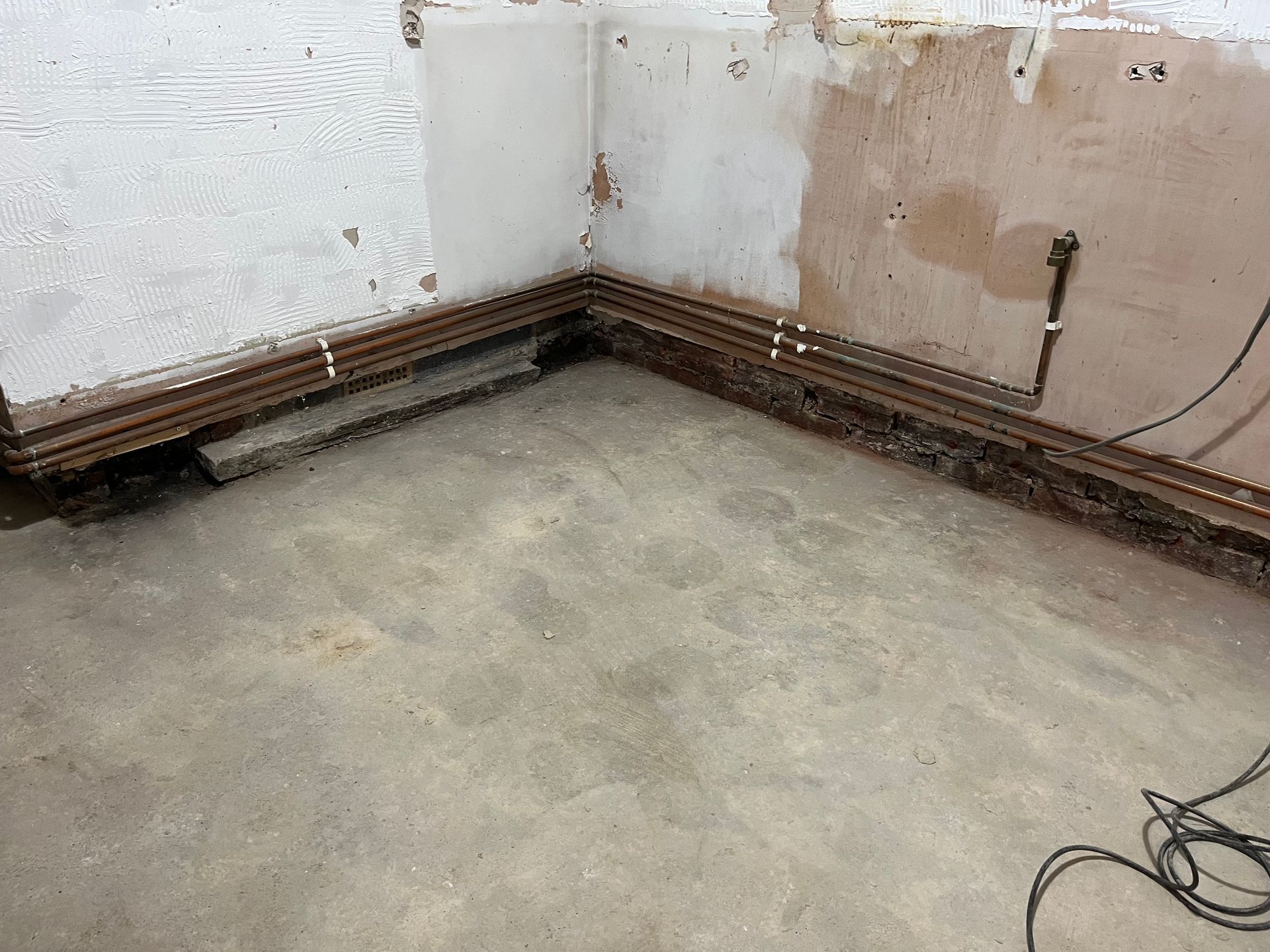 Corner of a room under renovation, bare concrete floor, exposed pipes along the walls, and damaged plaster.