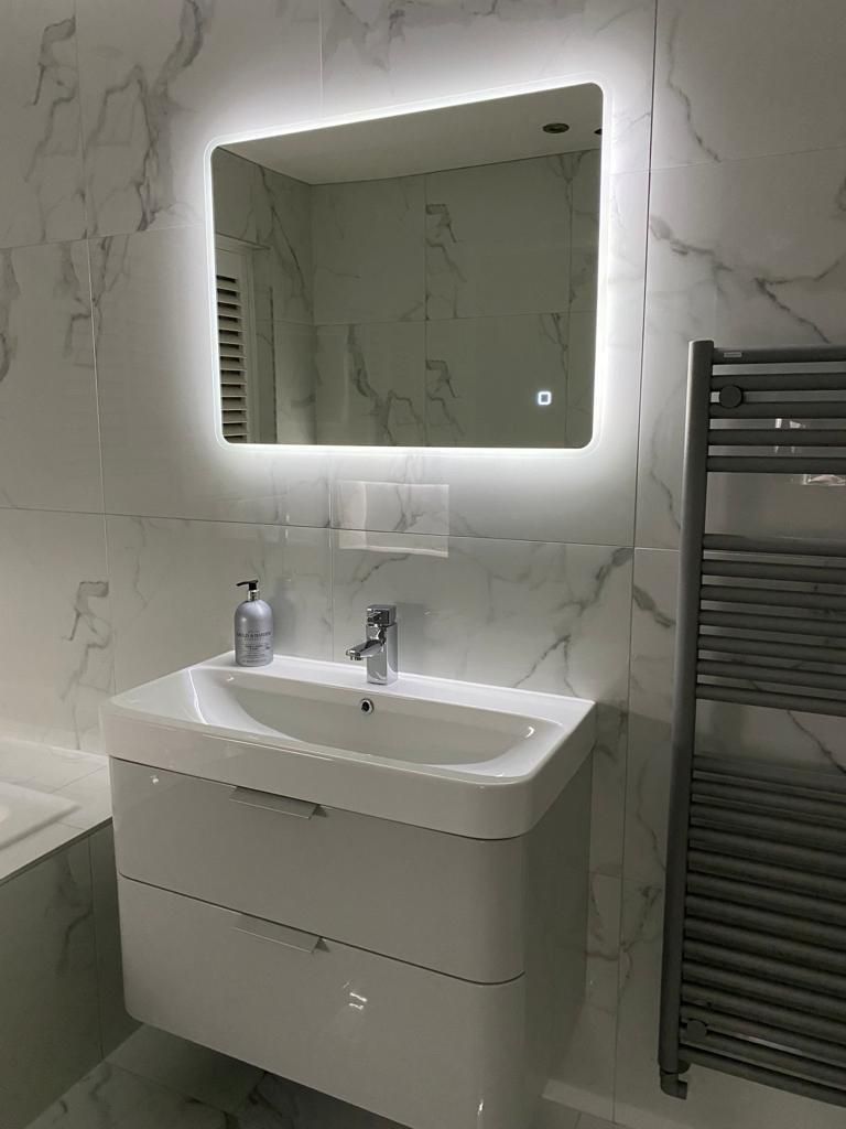 Modern bathroom with a white vanity, lighted mirror, and towel warmer against marble tile.