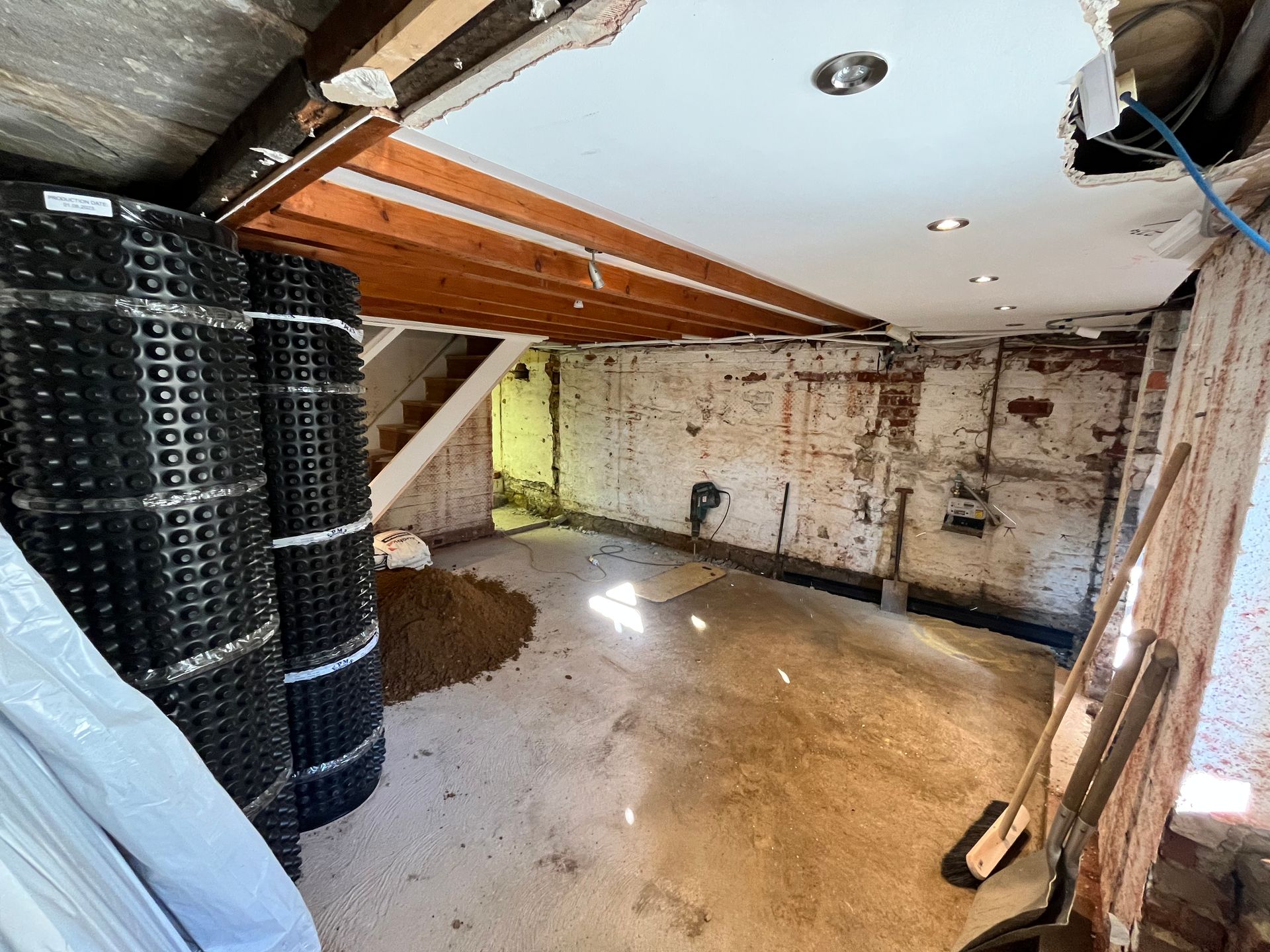 Basement renovation in progress with exposed brick, ceiling beams, and rolled black membrane; tools on the right.