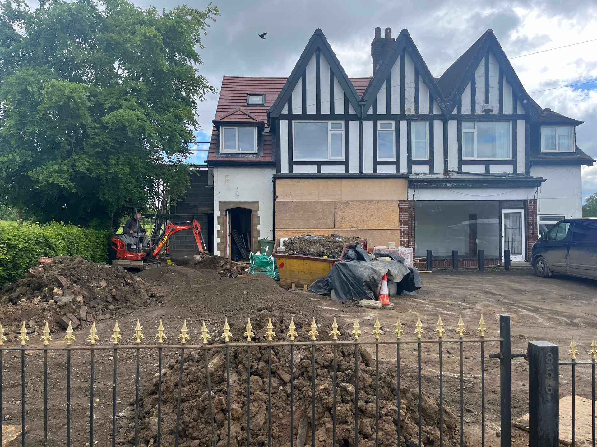 House under construction; facade with black and white timbering, dirt piles, excavator, and metal fence.
