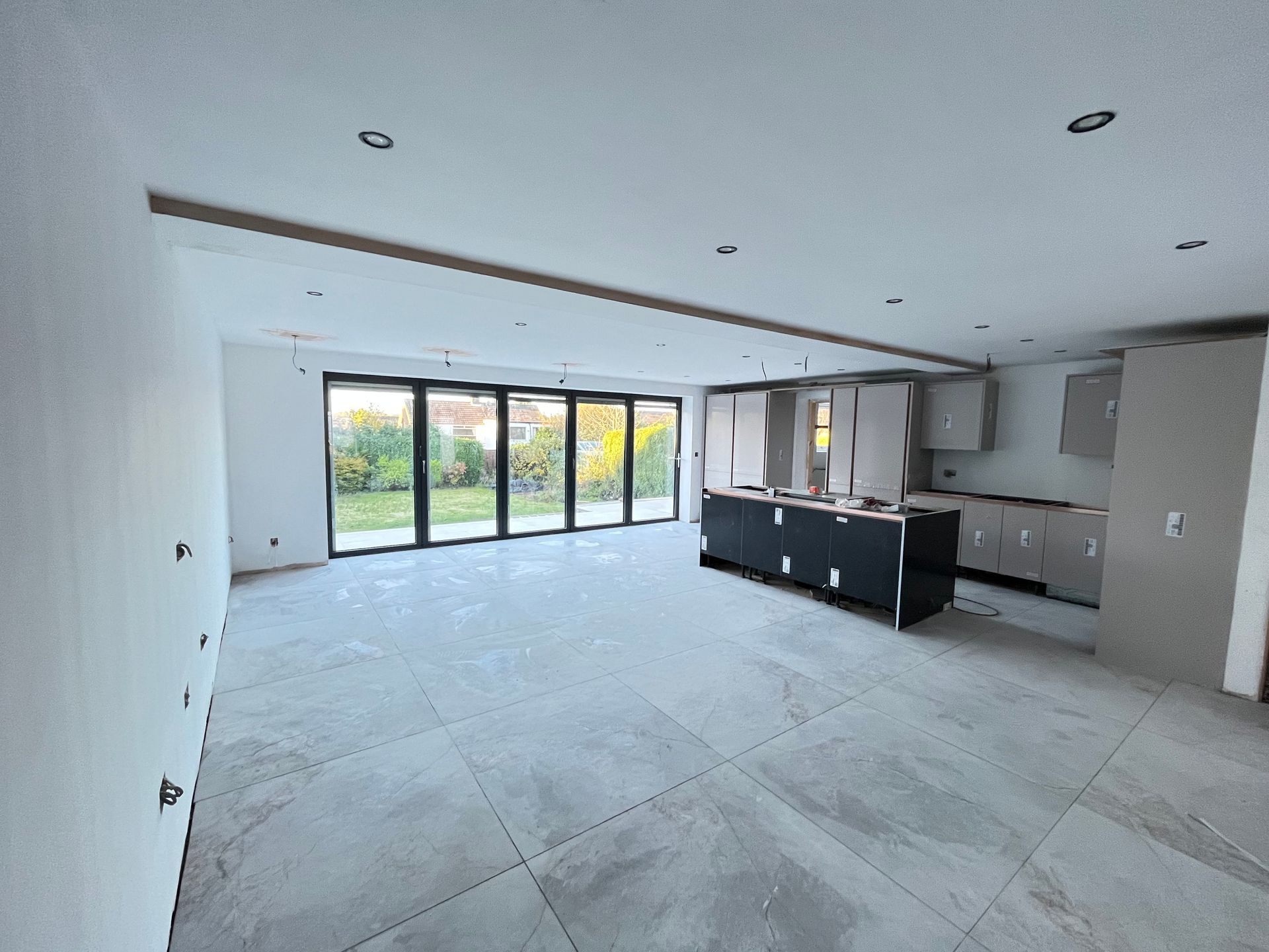 Open-plan kitchen under construction. Large windows lead to a yard, gray tile flooring, cabinets, and a black island.