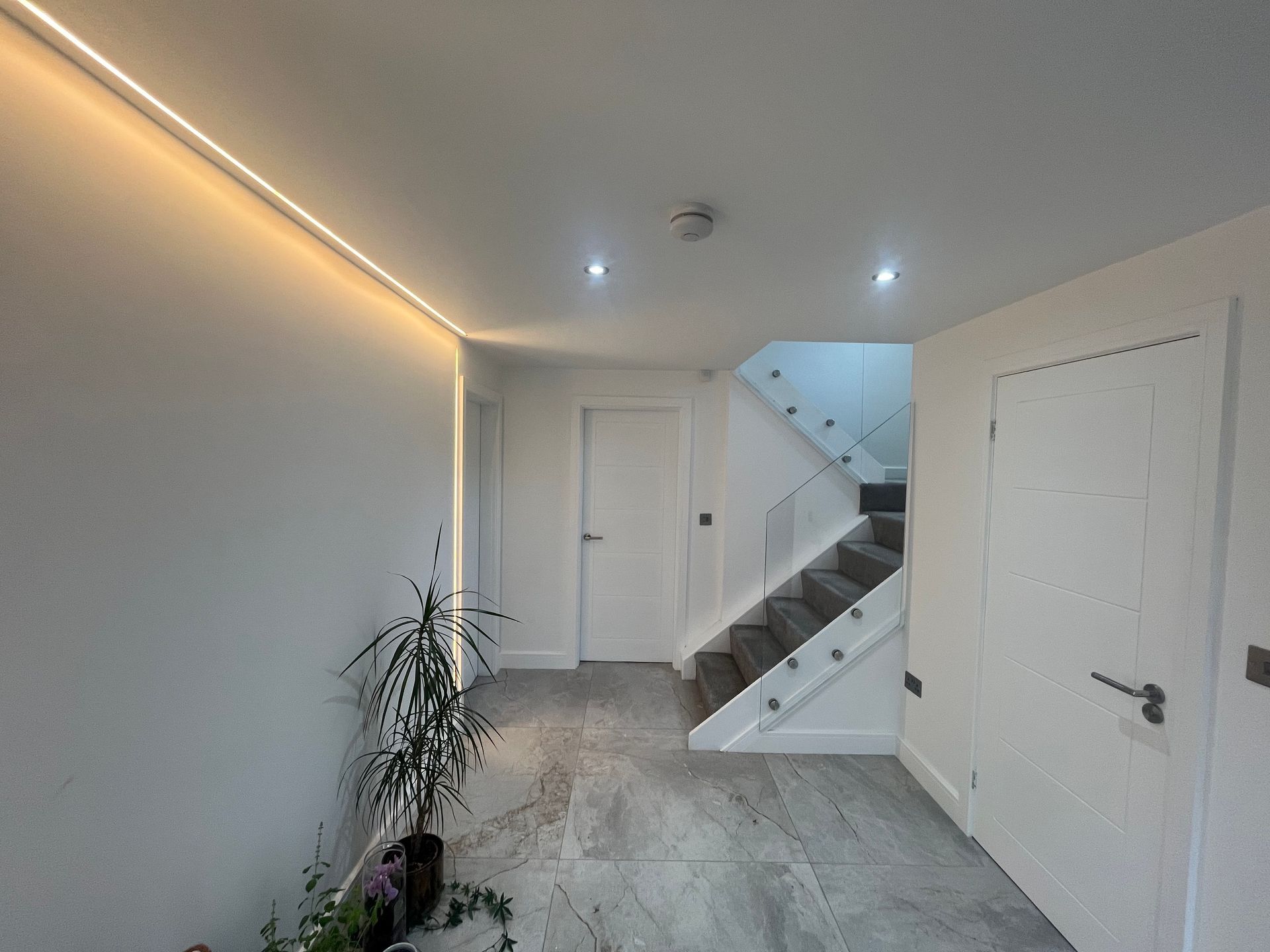 White hallway with recessed lighting, stairs, and a plant.