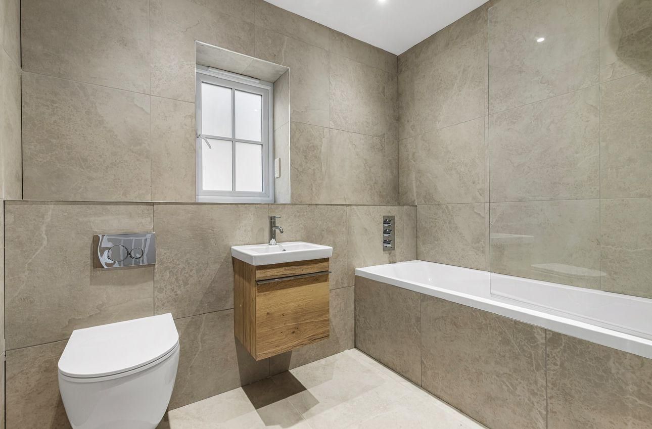 Modern bathroom with beige tile, toilet, wooden vanity, and bathtub. Small window.