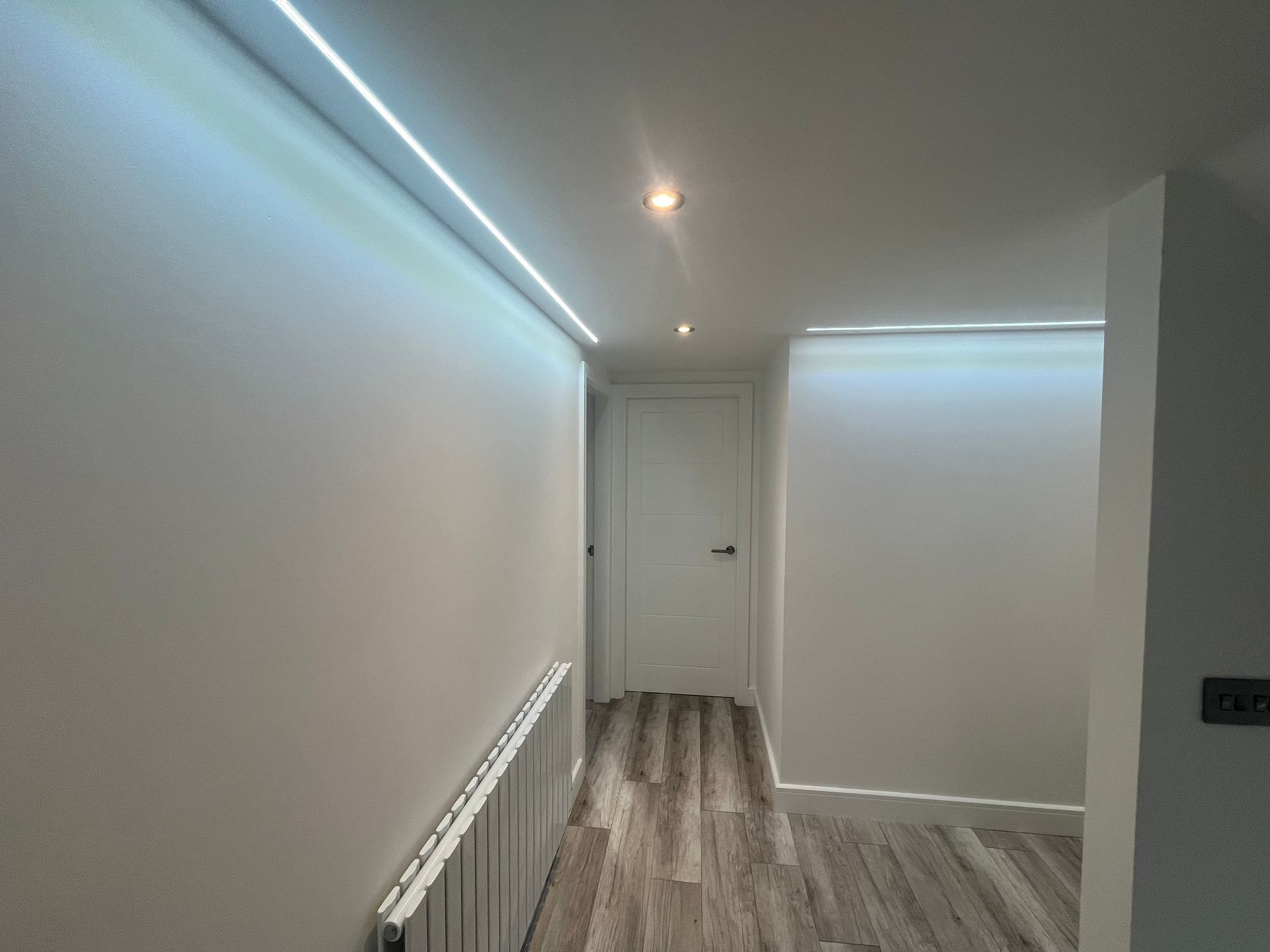 A modern hallway with recessed LED lighting, white walls, a radiator, and wood-look flooring.