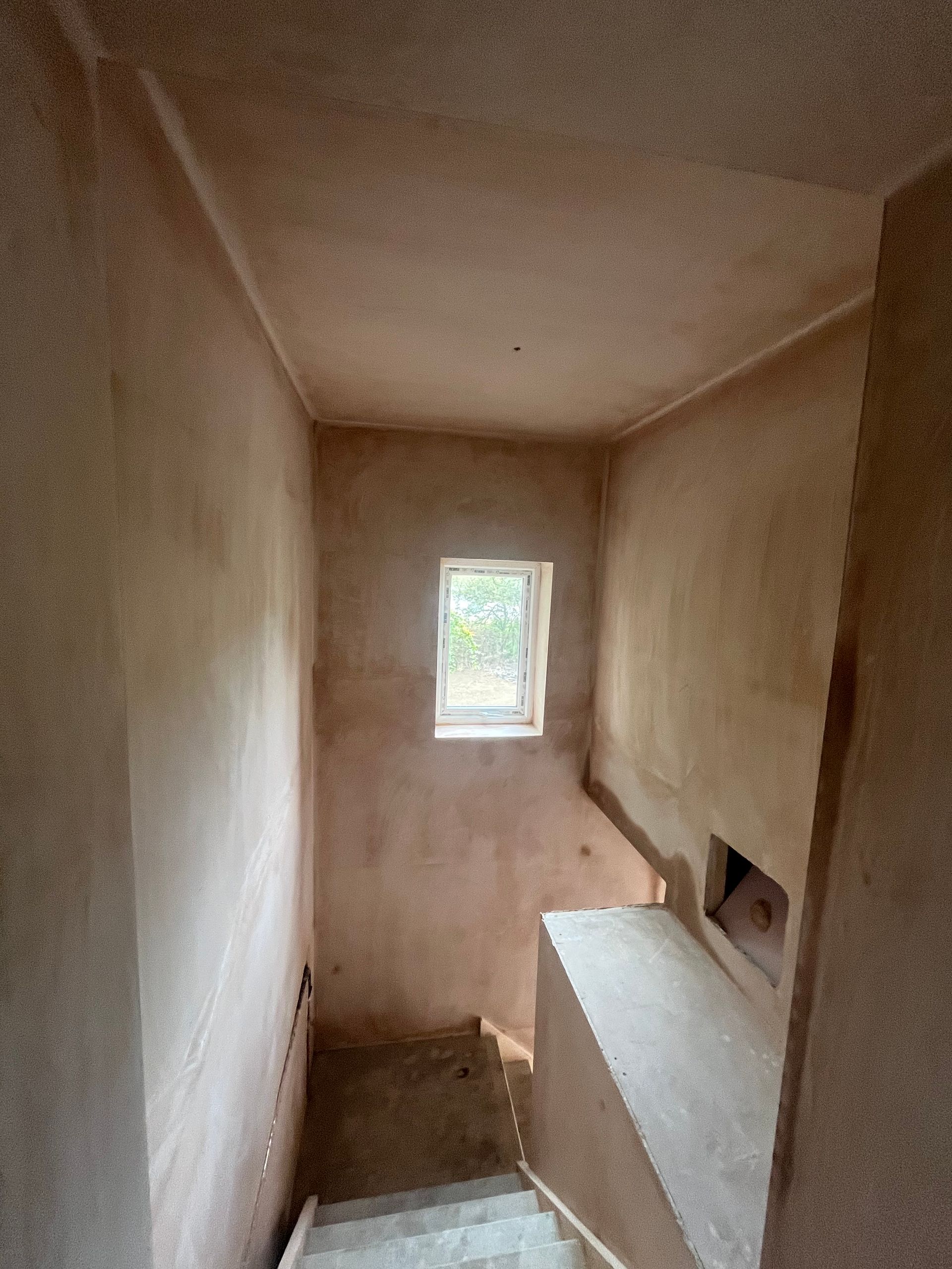 Stairwell with unfinished plaster walls and small window. Dusty tan color. Stairs lead down.