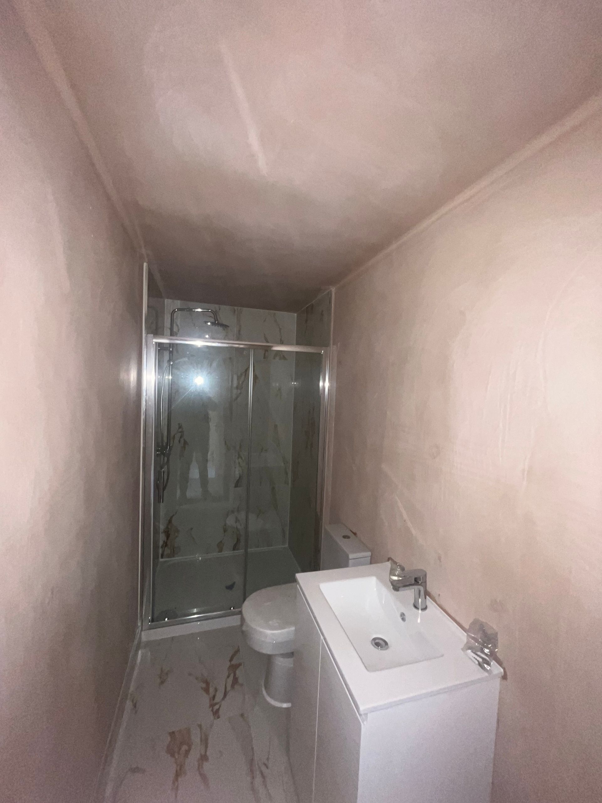 Narrow bathroom with shower, toilet, and sink, all against lightly pink plastered walls and a textured tile floor.