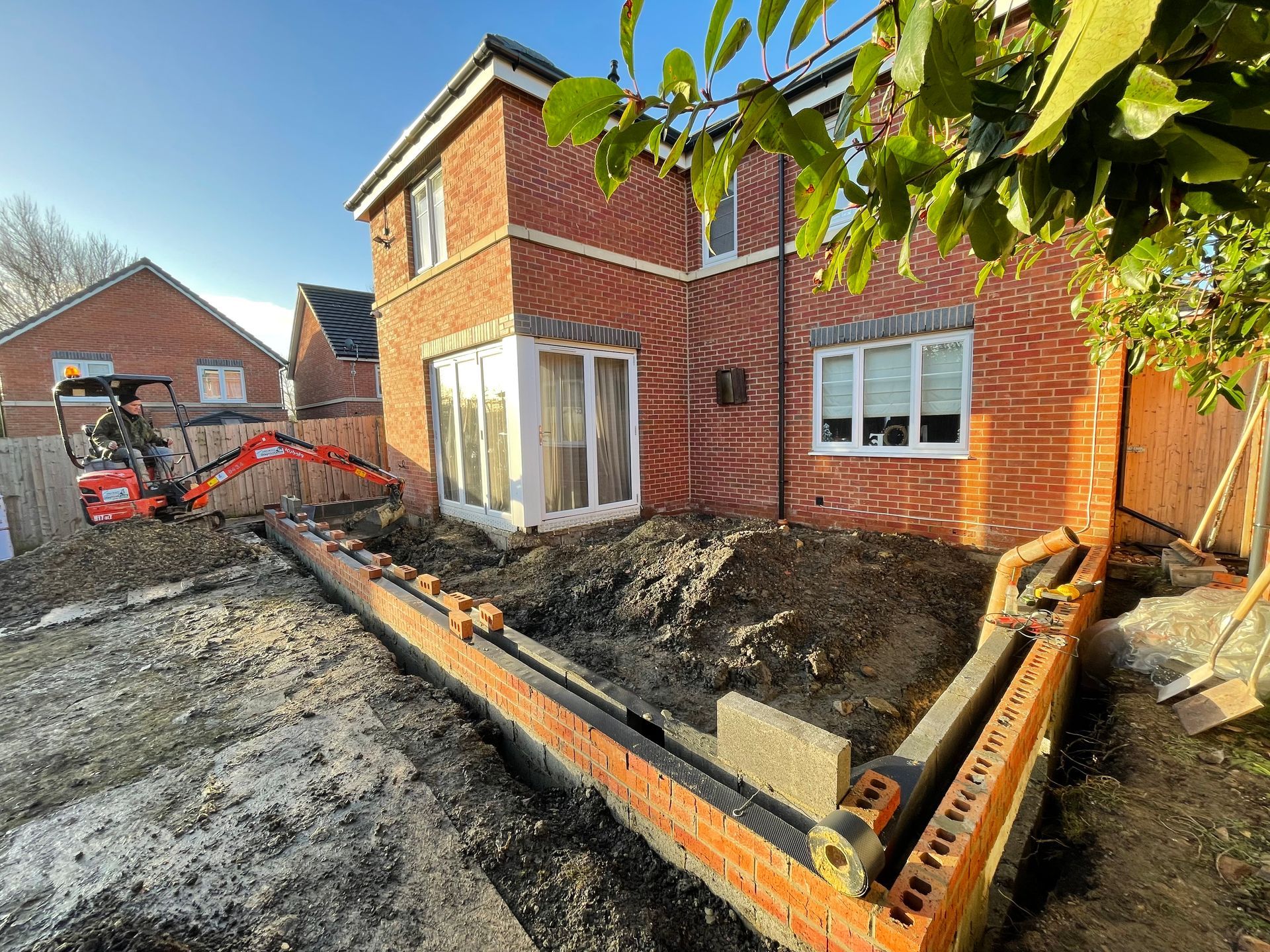 Brick retaining wall under construction near a red brick house, with a small excavator and a sunny setting.