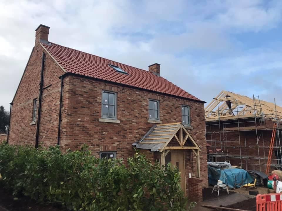 Brick house under construction with red roof and light blue sky. Another house under construction in background.