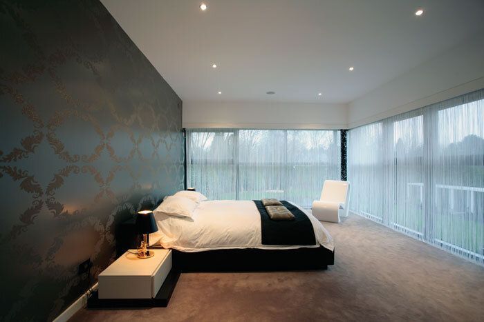 Bedroom with patterned wallpaper, bed with black blanket, white chair, and sheer curtains.