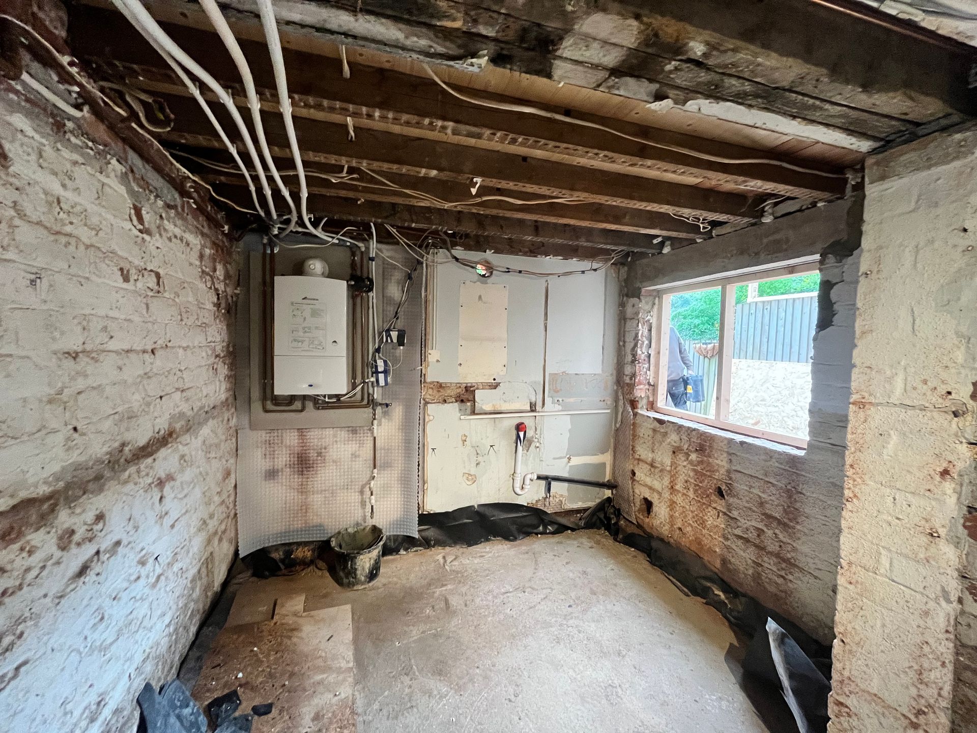 Interior of a room under construction: exposed brick walls, visible ceiling beams, window, boiler, and electrical wiring.