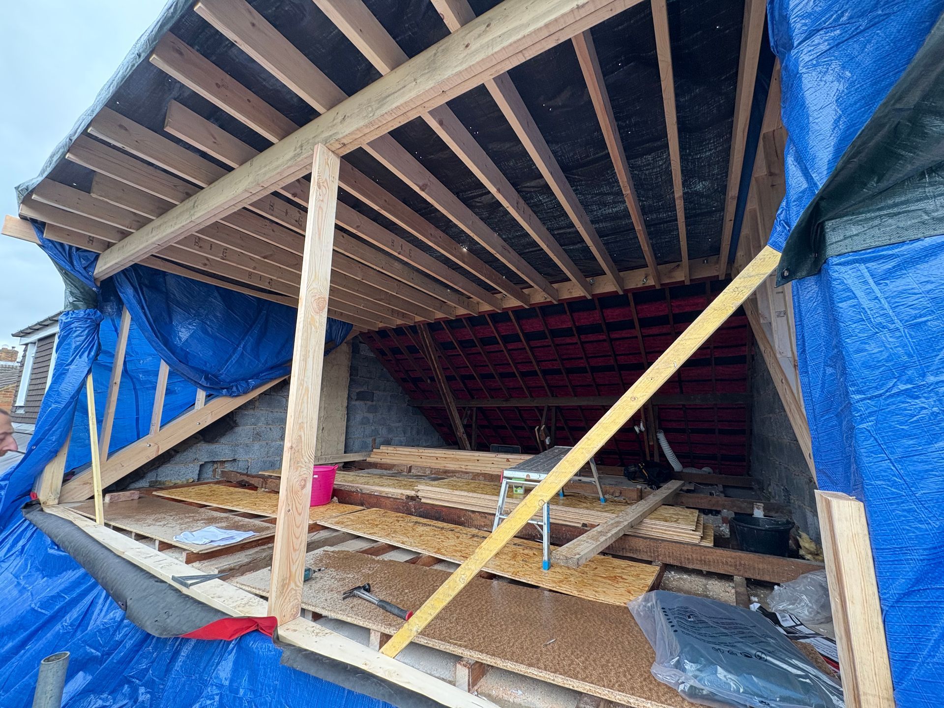 Construction site: a partially built roof with wooden beams, blue tarp, and materials inside.
