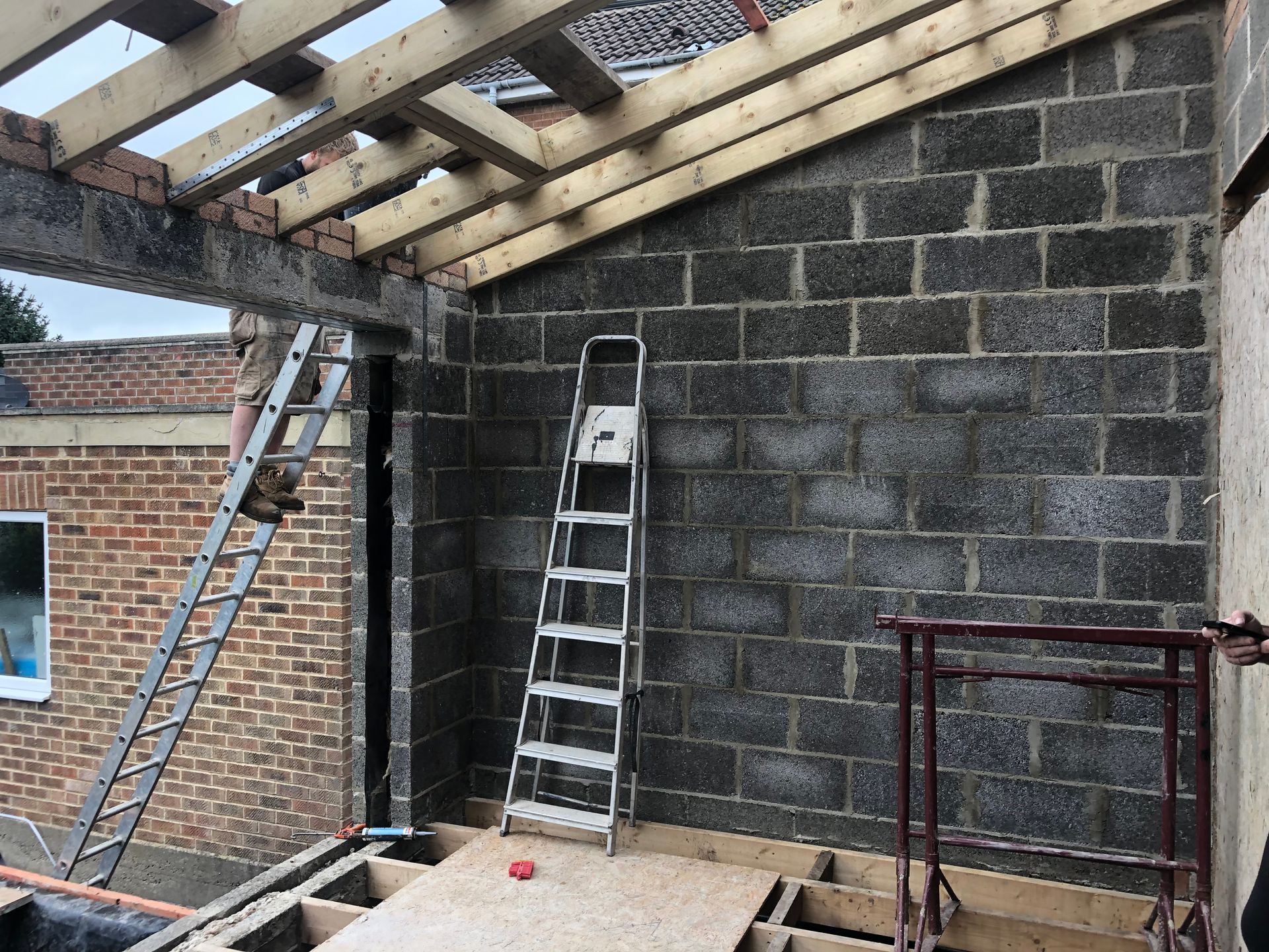 Construction site: concrete block wall, wood framing for roof and floor. Ladder and brick building visible.