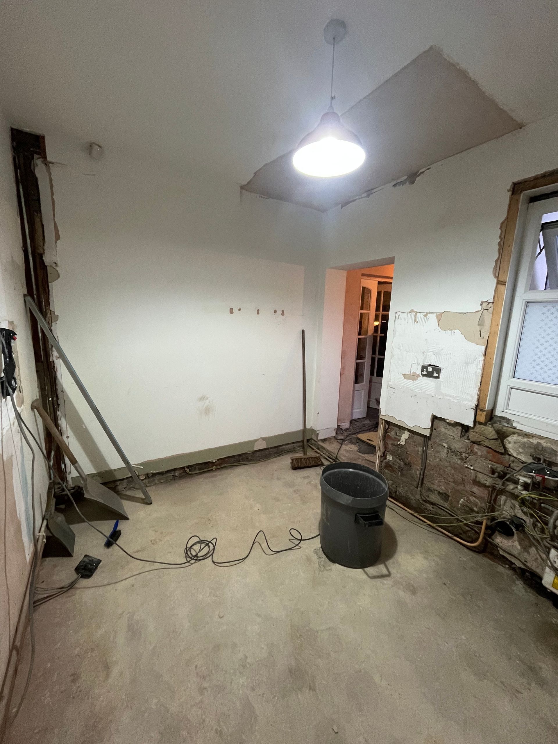 Room under renovation: walls stripped, exposed brick, debris on floor, overhead light.