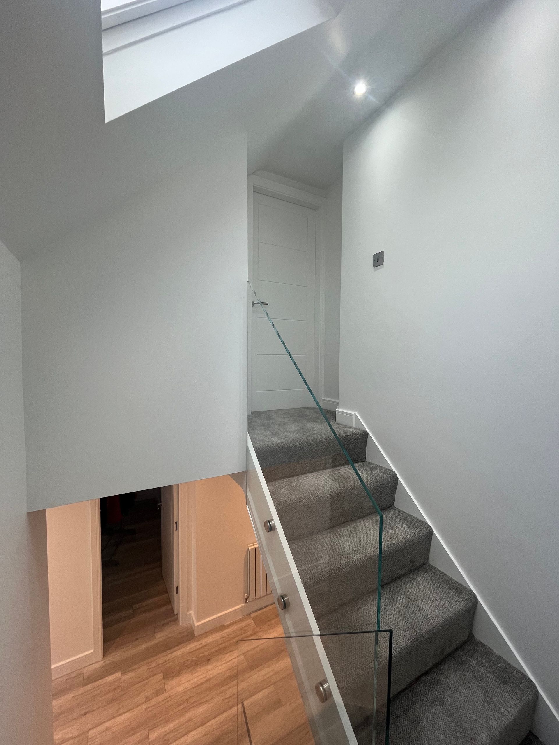 Staircase with glass railing and gray carpet. Sunlight enters from skylight. Entryway visible below.