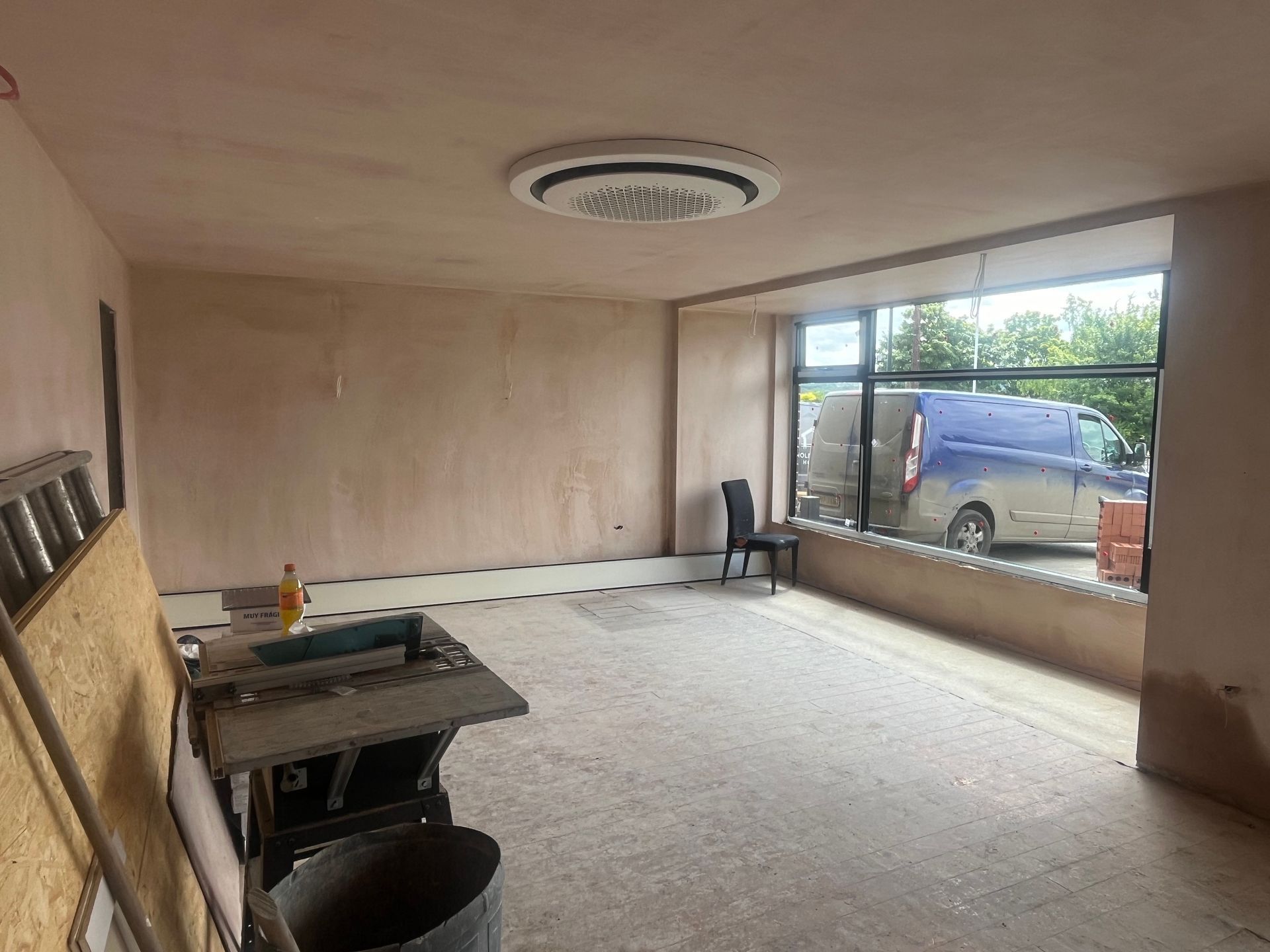 Empty room with fresh plaster, circular ceiling vent, large window with a van parked outside.