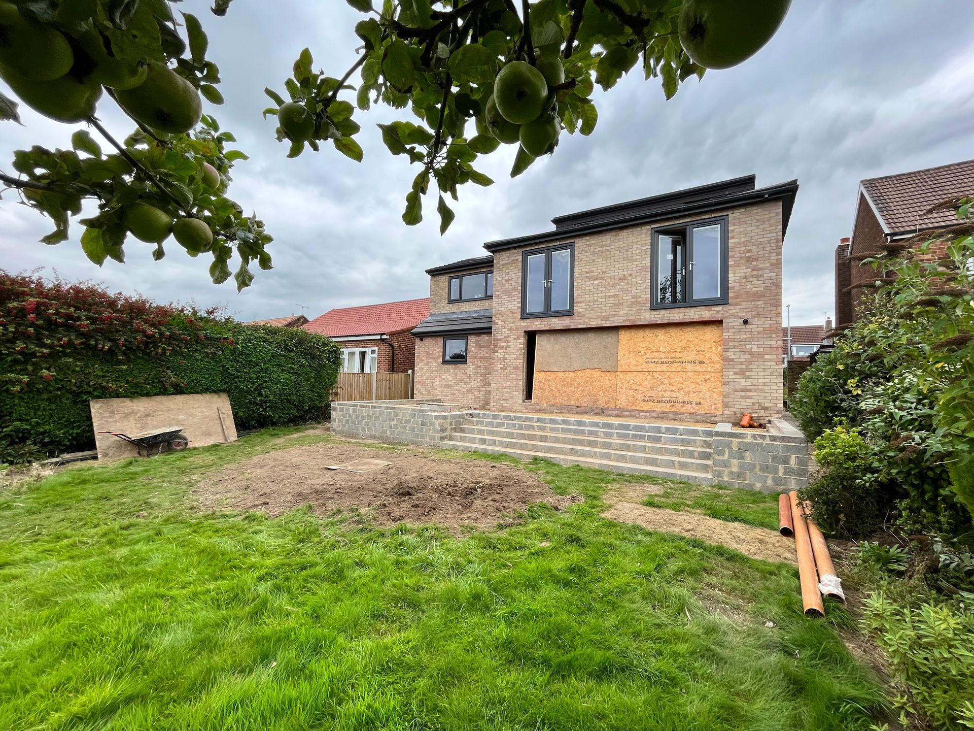 Backyard with a two-story brick house under construction. Cloudy sky, apple tree, and green lawn.