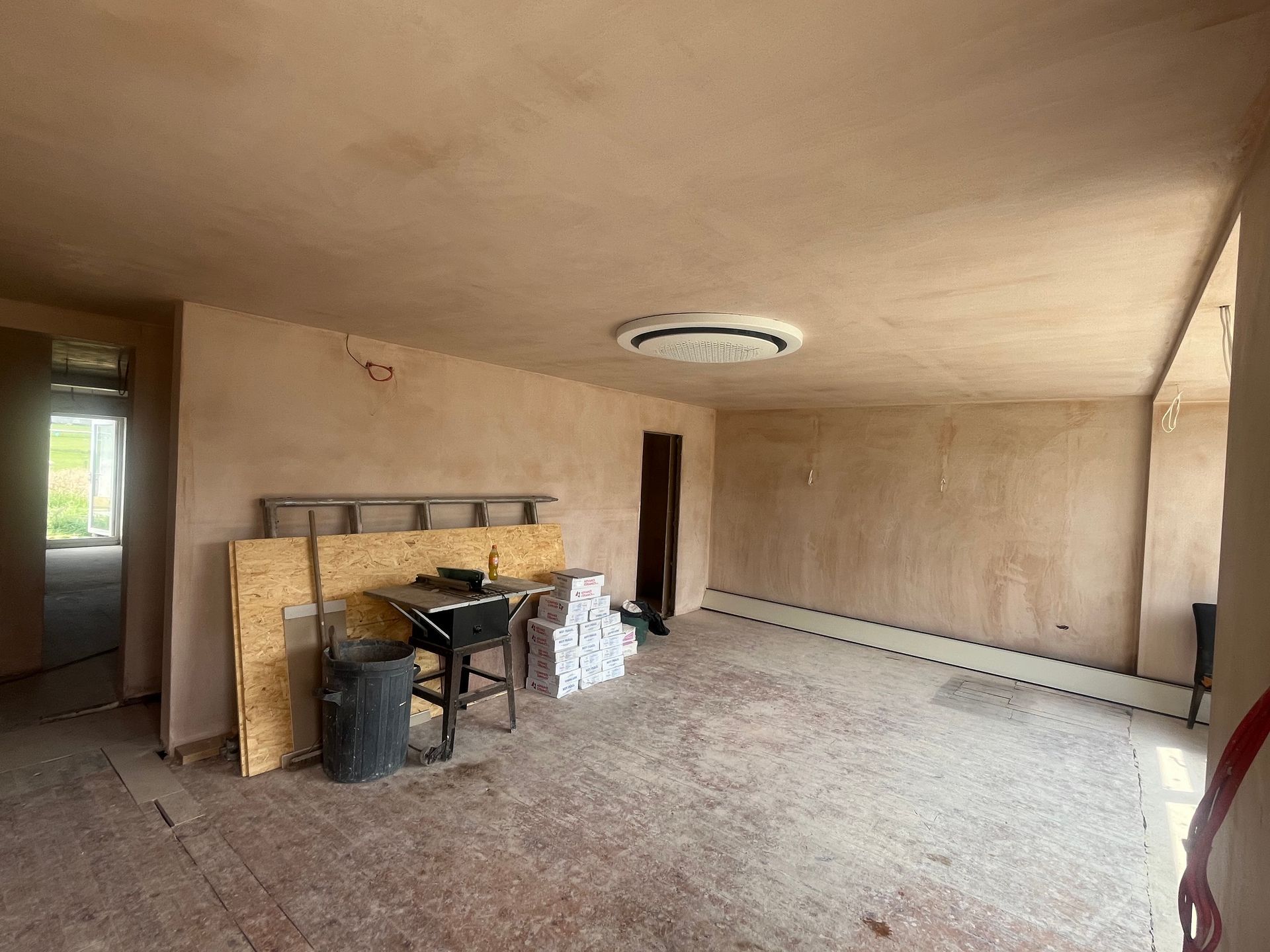 Empty room under construction; walls and ceiling plastered, unfinished wood floor, tools, and materials scattered.