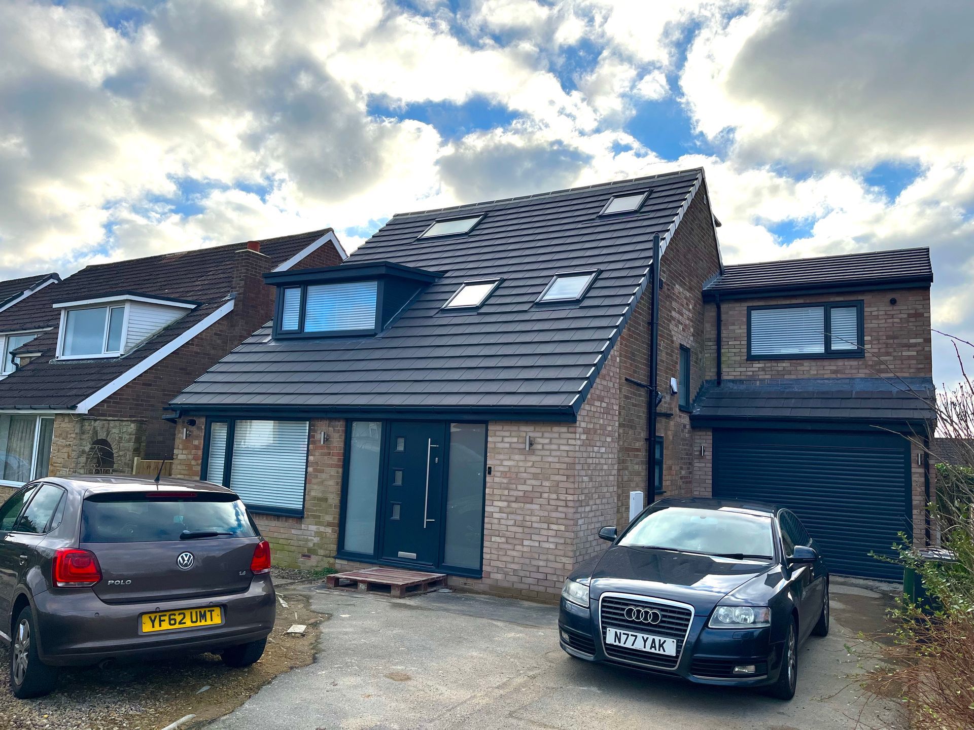 Modern brick house with dark roof, garage, and two cars parked in the driveway under a cloudy sky.