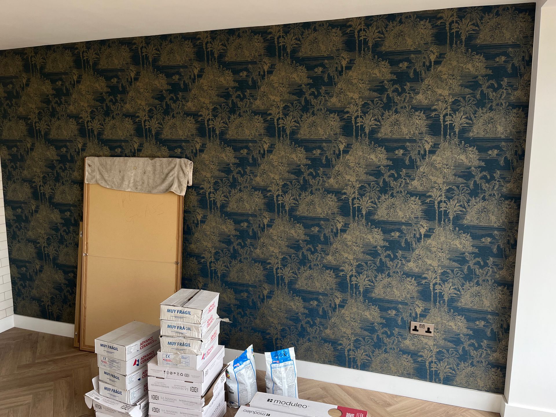 Room with teal and gold wallpaper, building materials, and a large, beige panel leaning against the wall.