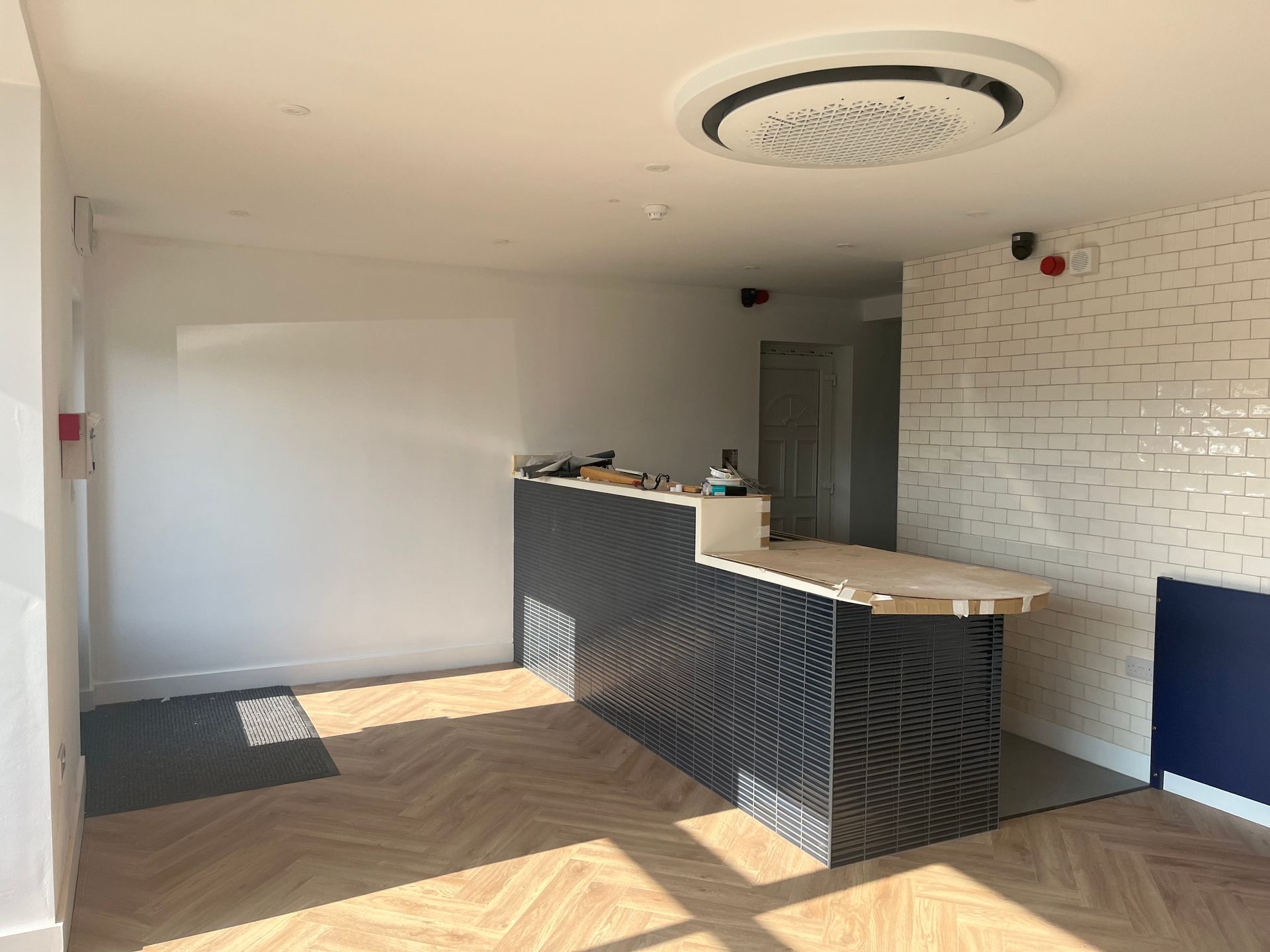 Empty modern office reception area with a patterned wooden floor, dark gray tiled counter, and white brick wall.