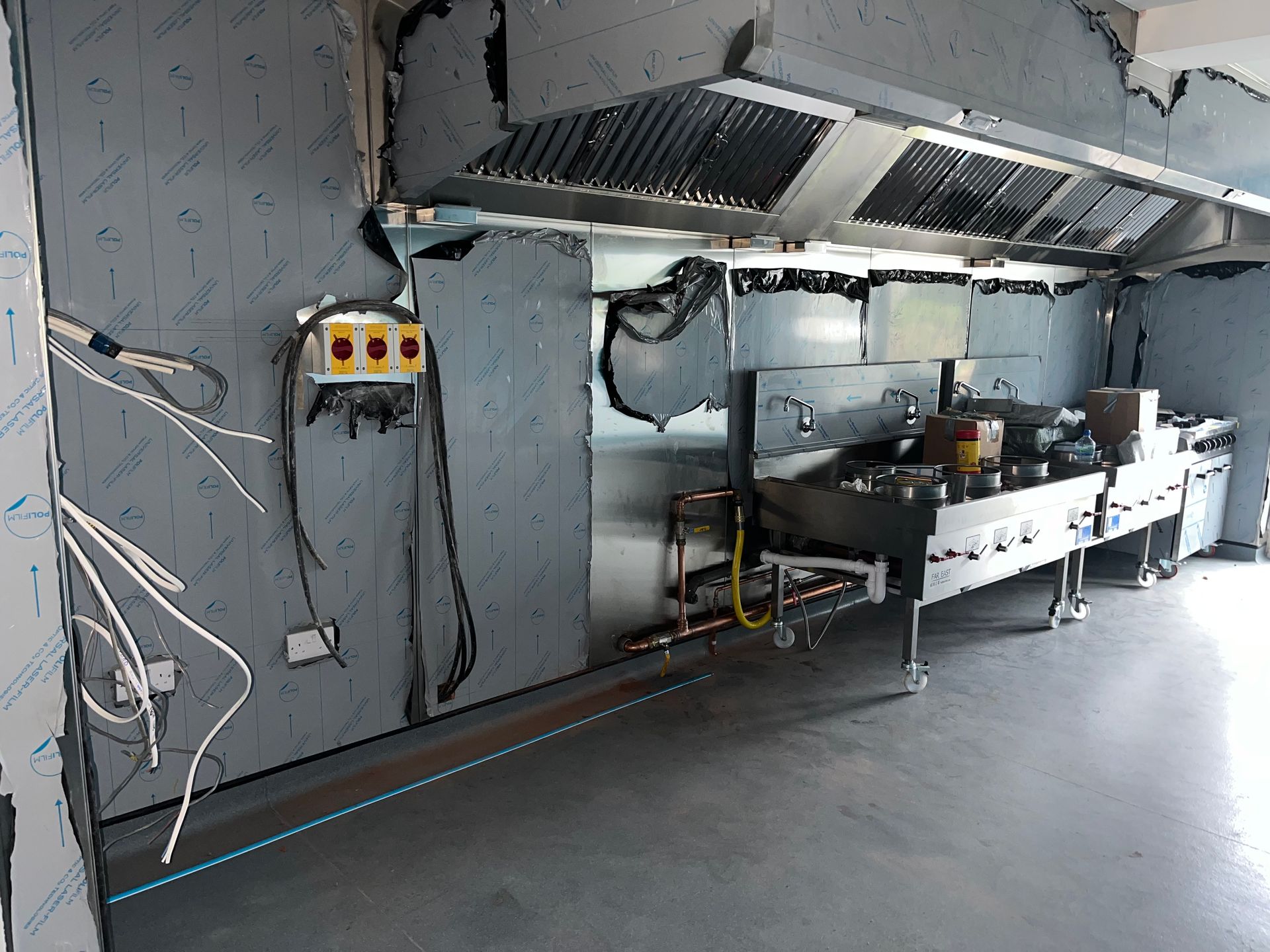 Commercial kitchen with stainless steel equipment, ventilation hood, control panel, and exposed wiring.