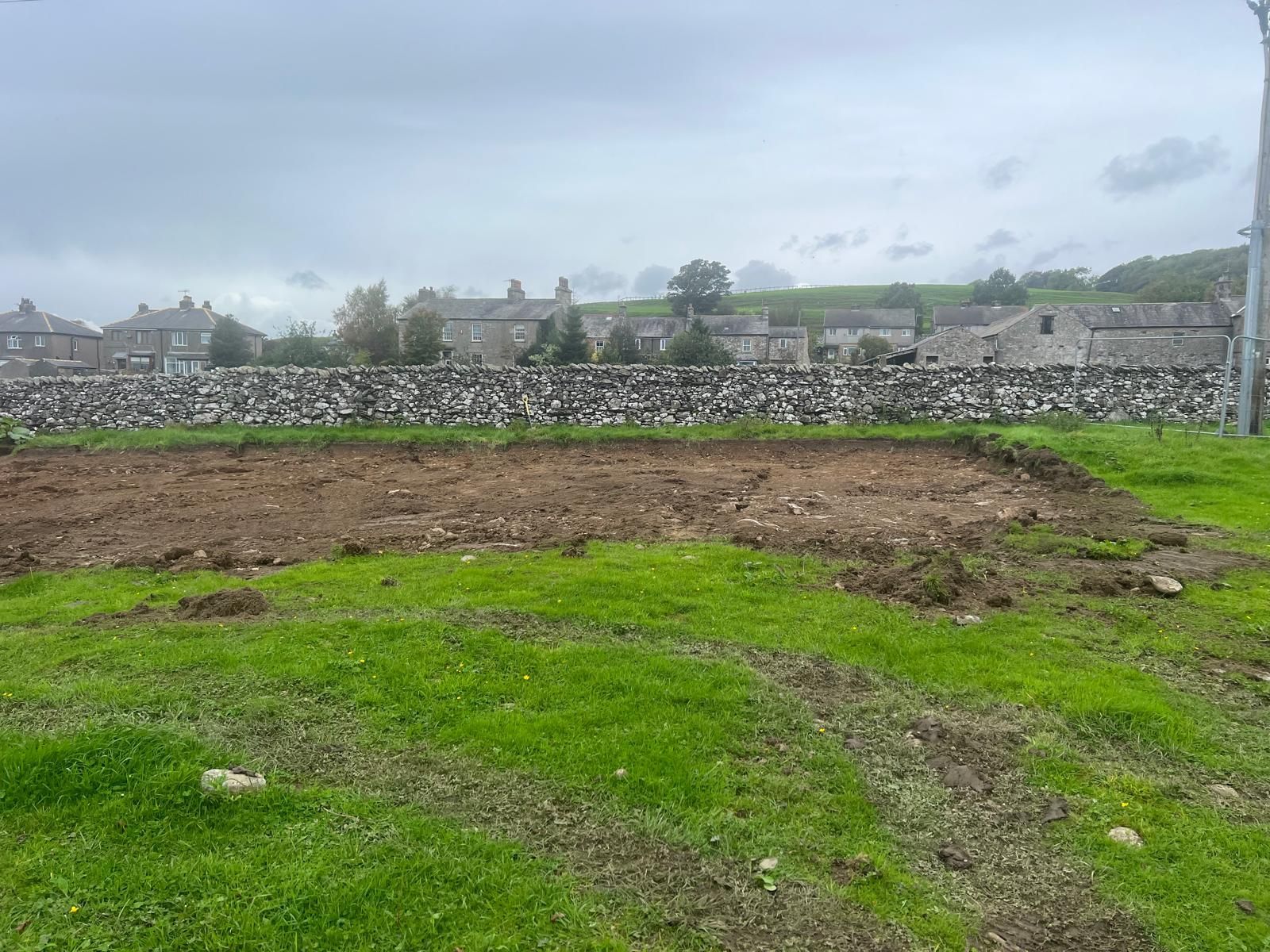 A field of green grass and tilled brown earth with a stone wall and buildings in the background under a cloudy sky.
