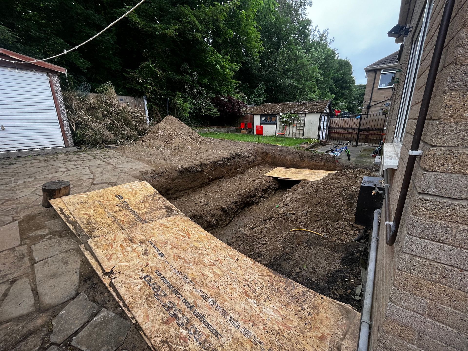 Backyard with excavated trenches, plywood, and piles of dirt near a house.