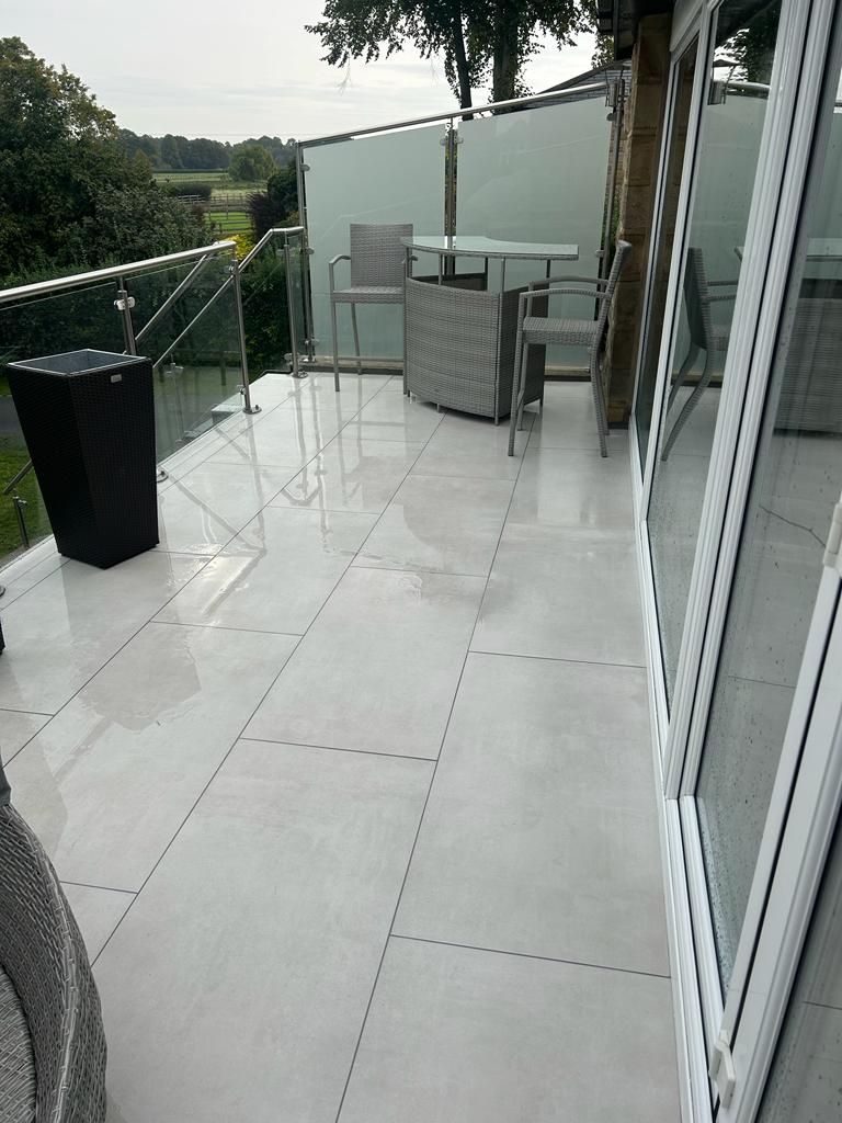 Bright patio with light-colored tiled floor, glass railing, and outdoor furniture; view of the yard beyond.