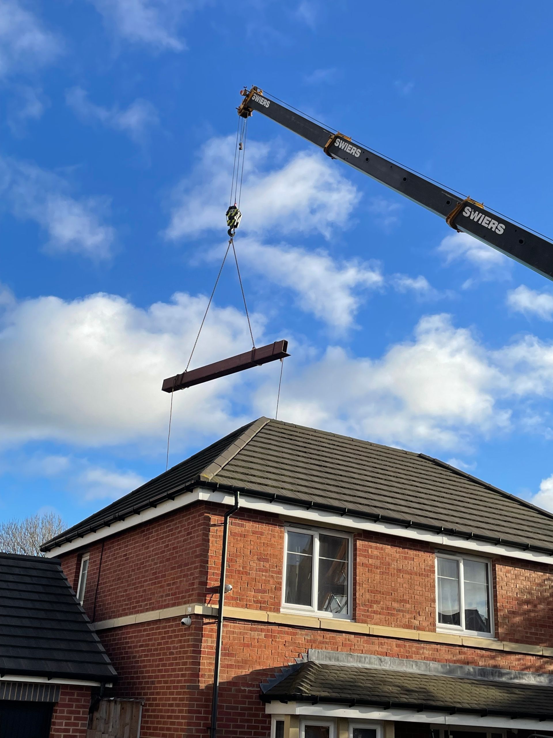 A crane lifting a long, brown beam over a brick house under a blue sky.