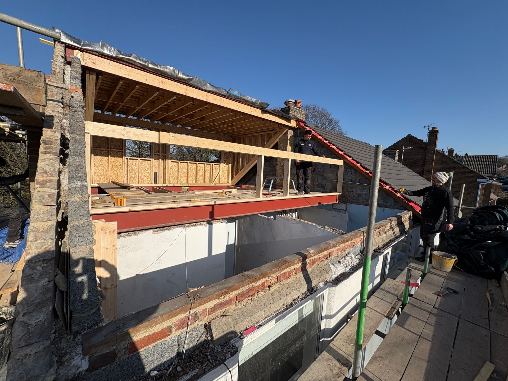 Construction site: workers building a roof addition, with a red steel beam and exposed wood framing.