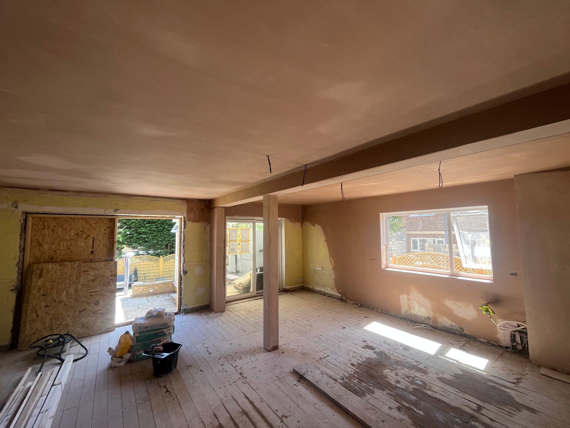 Interior of a room under construction with plastered walls and a wooden floor. Sunlight streams through windows.