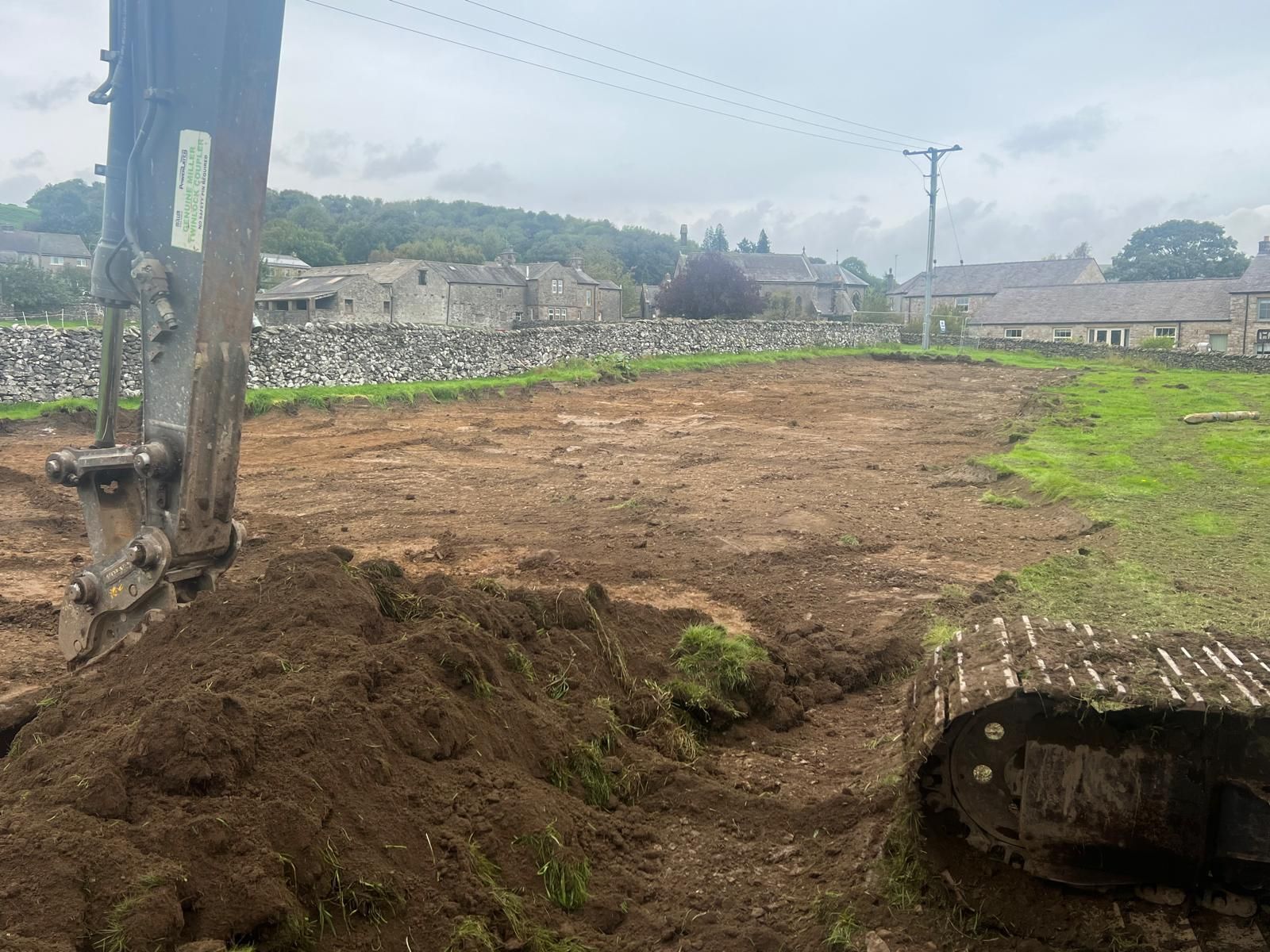 An excavator digging in a field, clearing the land. Buildings in the background under an overcast sky.